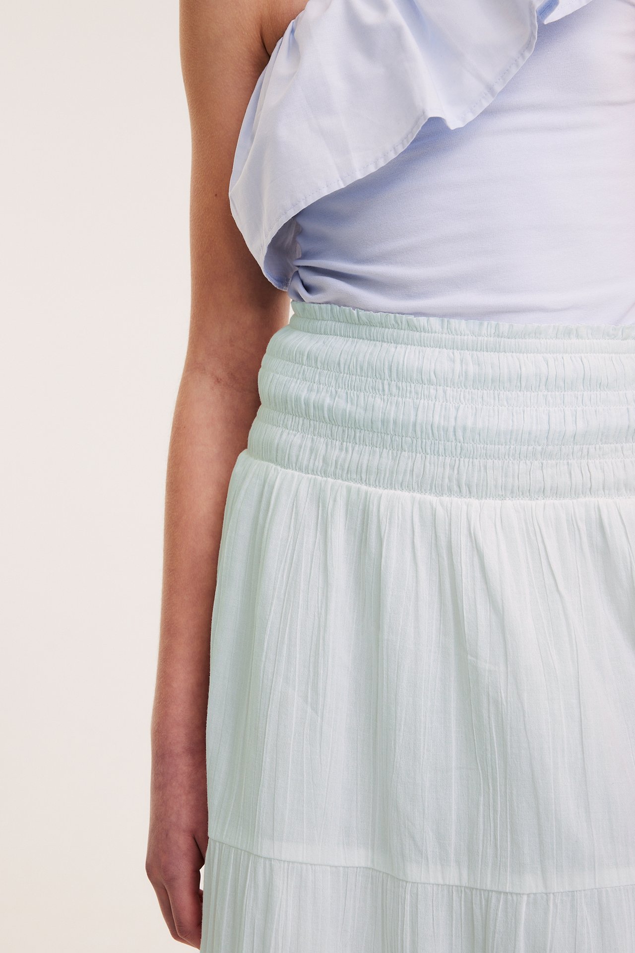 Kobieta w jasnozielonej marszczonej spódnicy maxi i jasnoniebieskim topie z falbaną, widok z przodu.