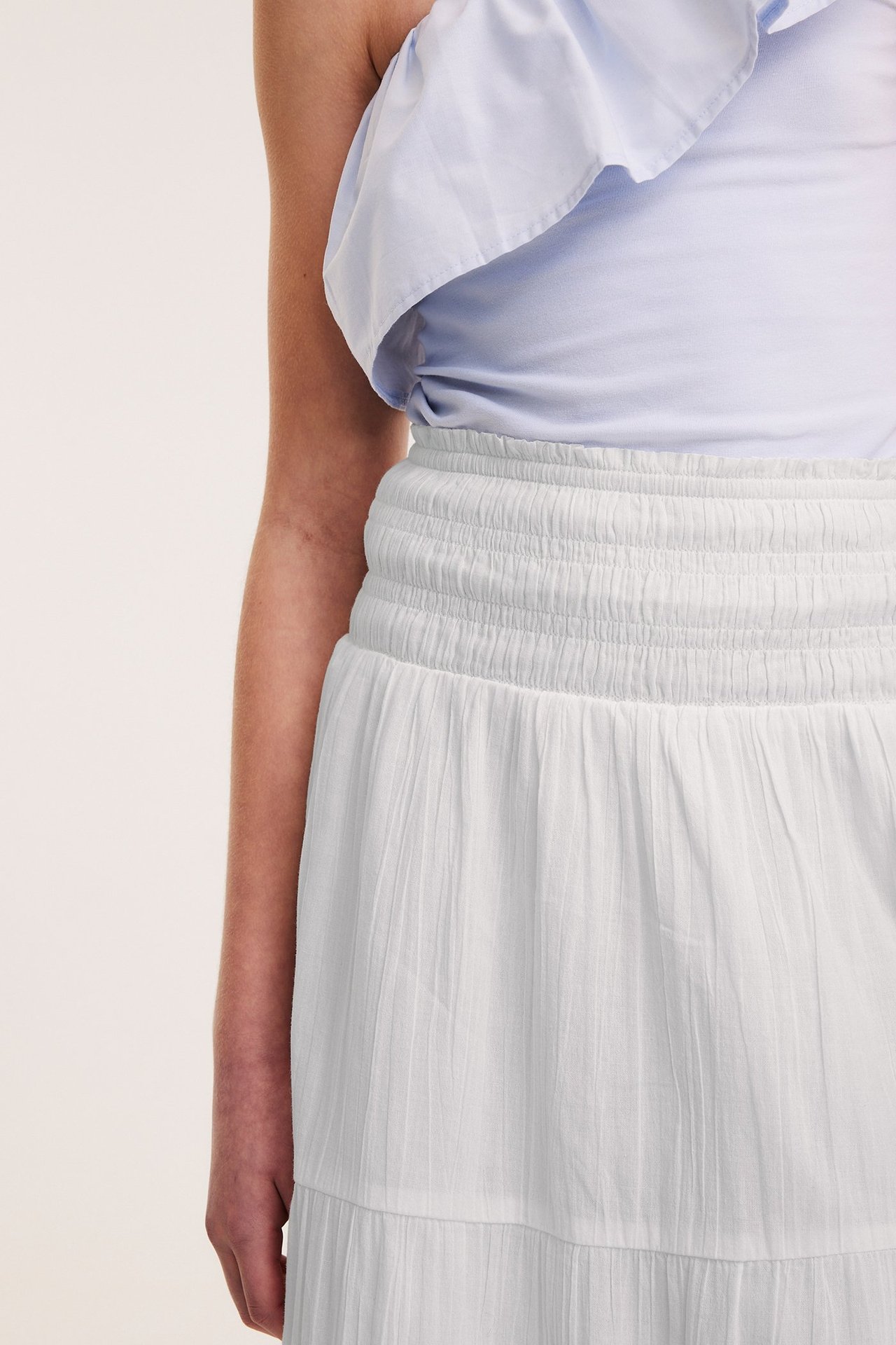 Zbliżenie kobiety w białej marszczonej spódnicy maxi i jasnoniebieskim topie z odkrytymi ramionami.
