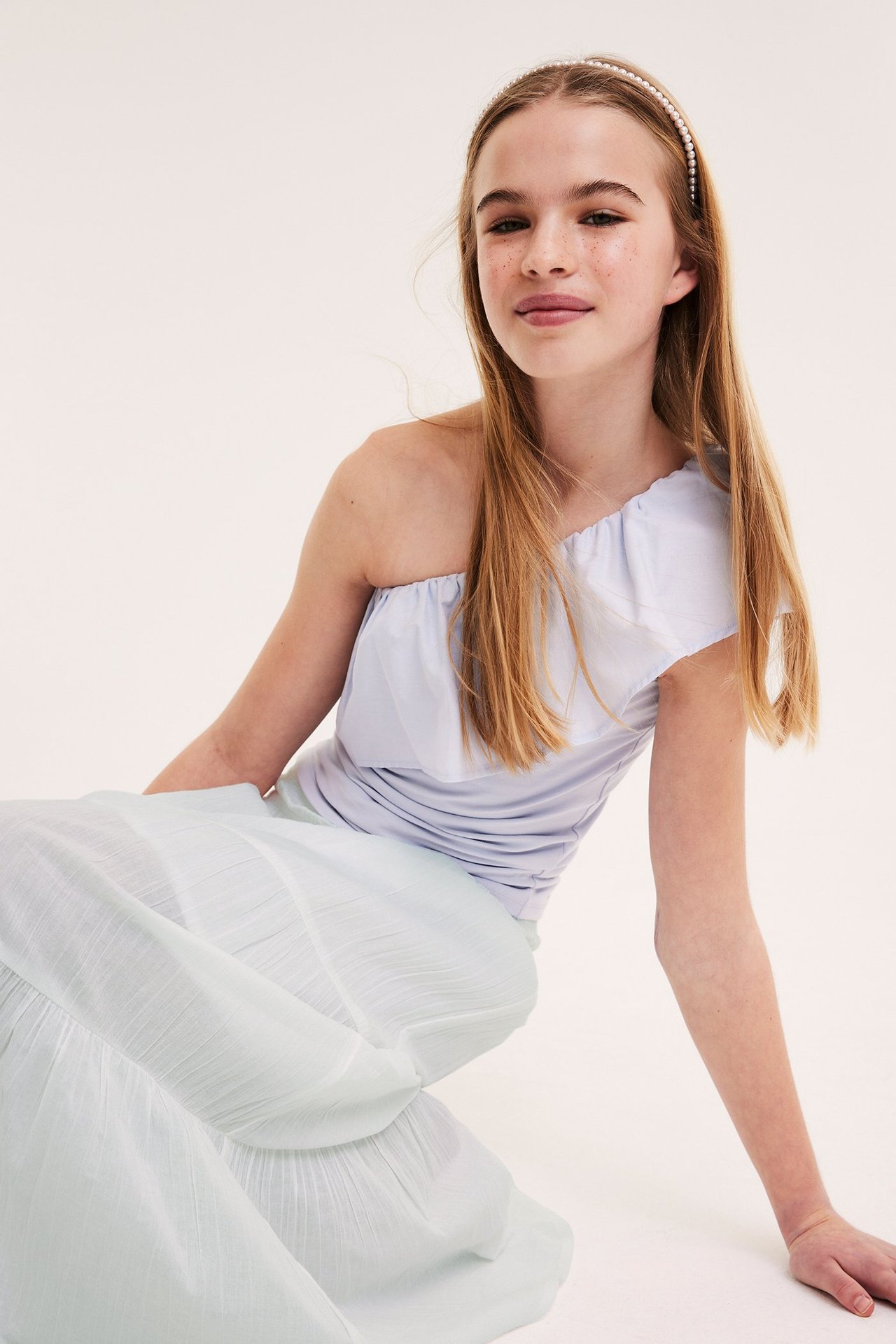 Widok z przodu: Dziecko w jasnozielonej spódnicy maxi z falbanami, jasnoniebieskim topie na jedno ramię i opasce z perełek.