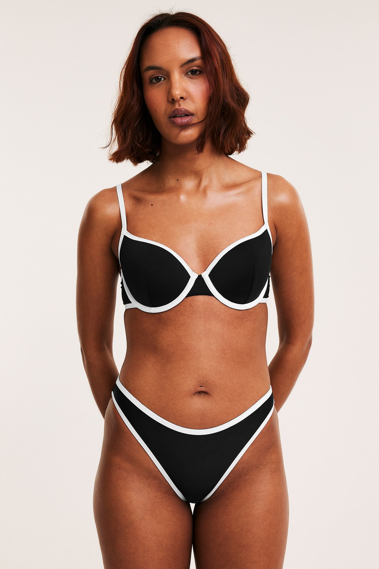 Frontvy: Kvinna i svart bikini-bh med vit kant och matchande svarta bikiniunderdelar.