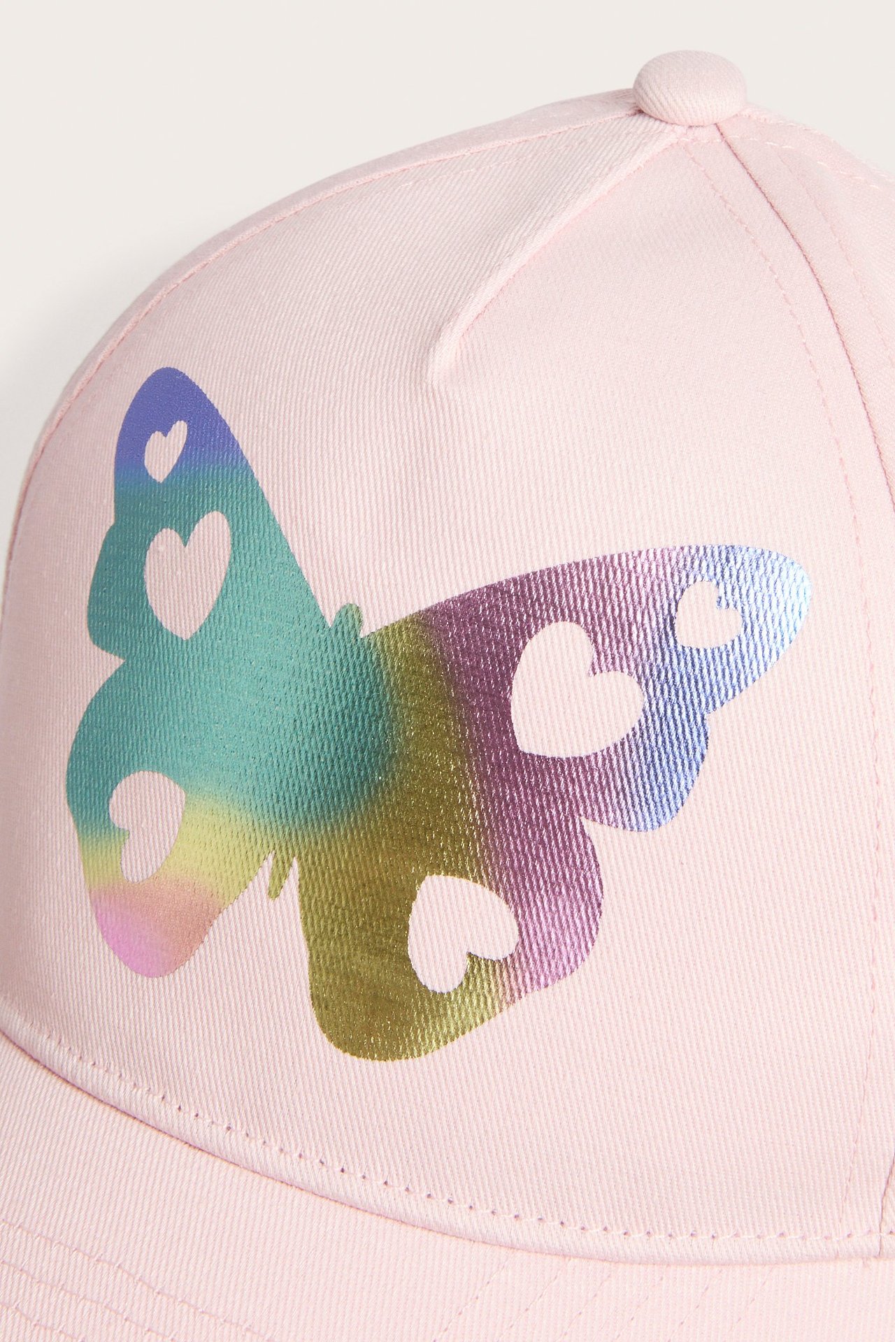 Zbliżenie różowej czapki z daszkiem z metalicznym, opalizującym nadrukiem motyla z wycięciami serc.