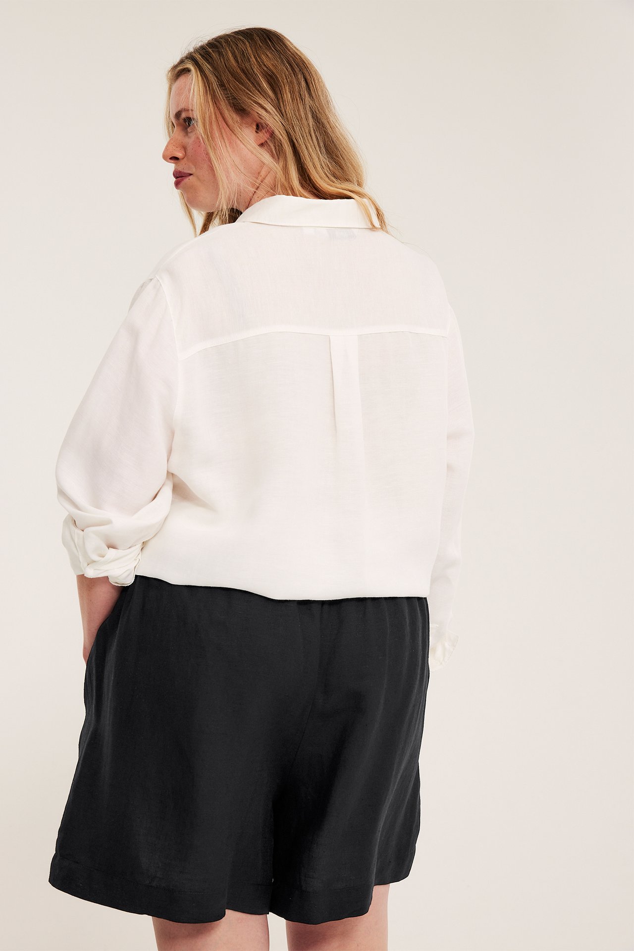 Nainen takaa/sivulta, yllään mustat pellava-viskoosishortsit smokkirypytyksellä ja valkoinen rento paita.