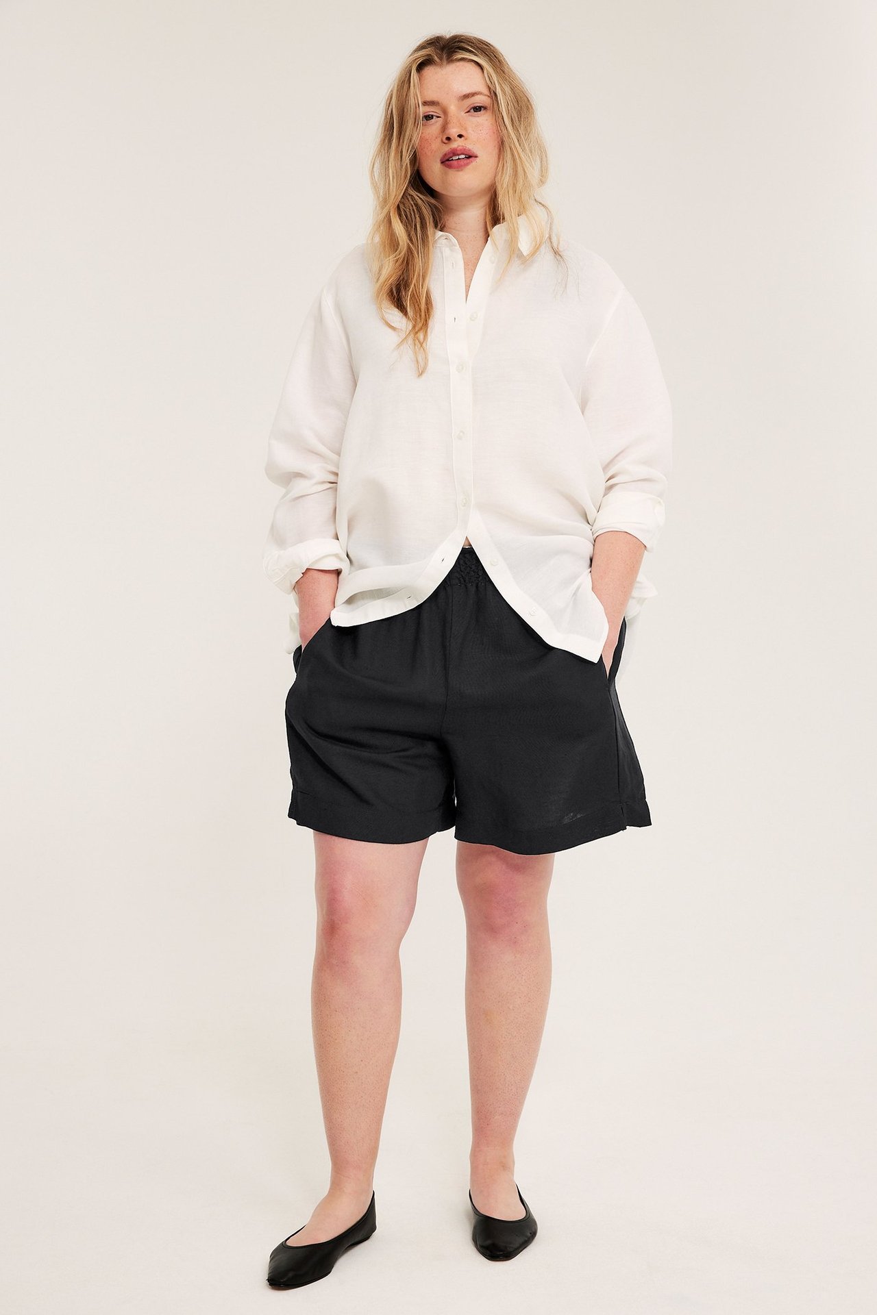 Forfra, kvinne i svarte viskose-linshorts med smock i midjen og en hvit linskjorte.