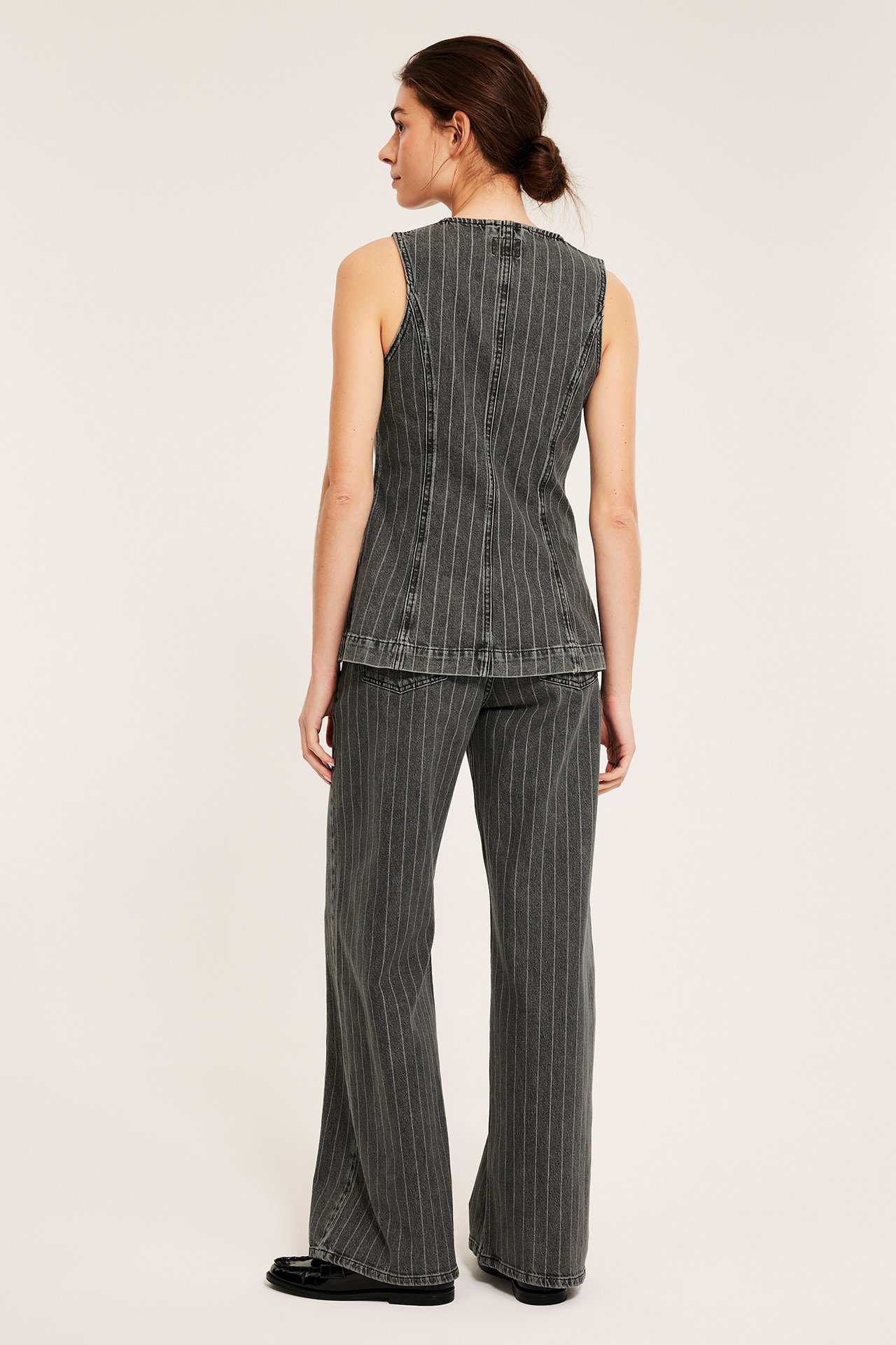 Bakre/sidovy av kvinna i mörkgrå kritstrecksrandiga jeans, matchande ärmlös topp och svarta loafers.