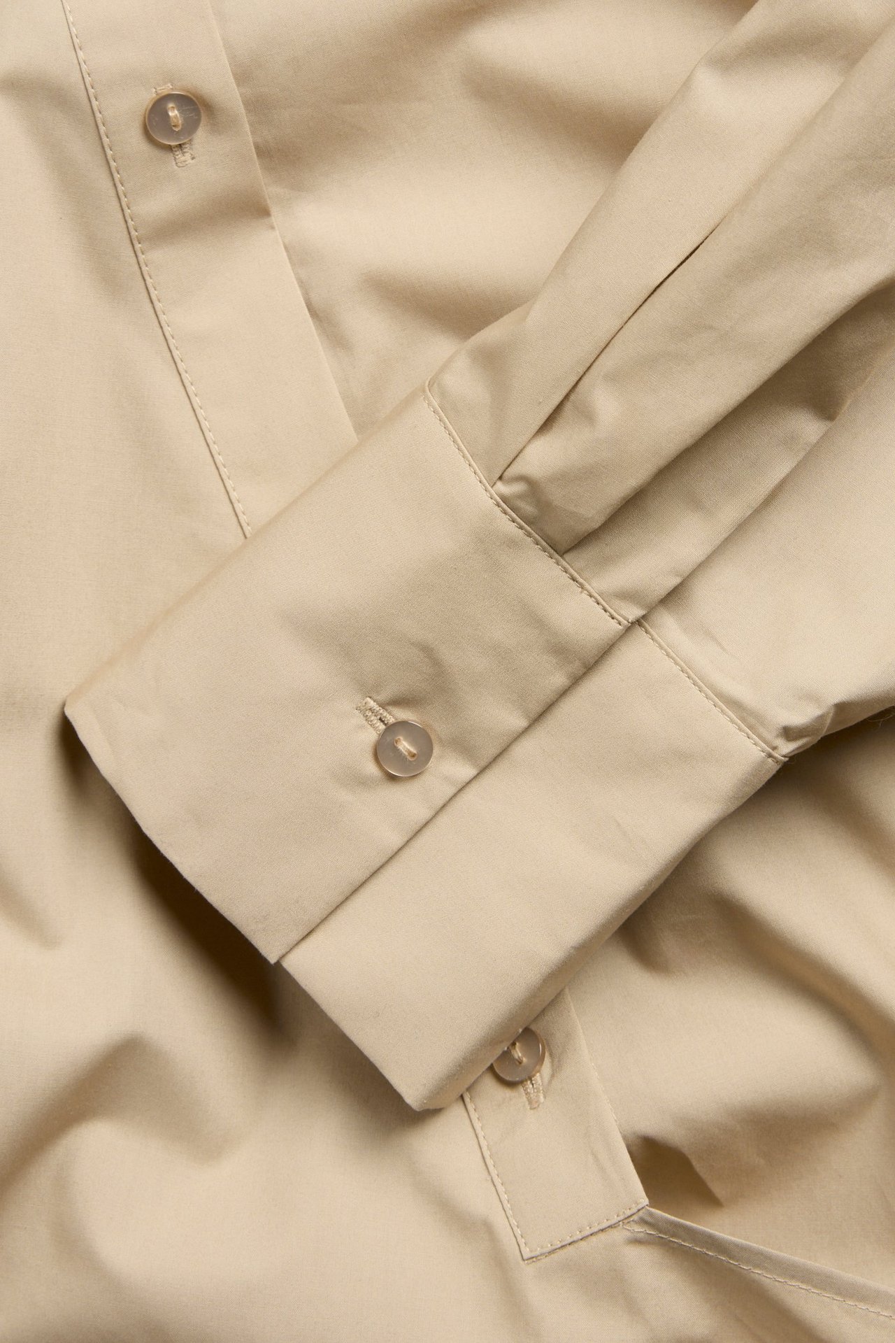 Zbliżenie beżowej koszuli bawełnianej, ukazujące mankiet z guzikiem i przednią plisę.