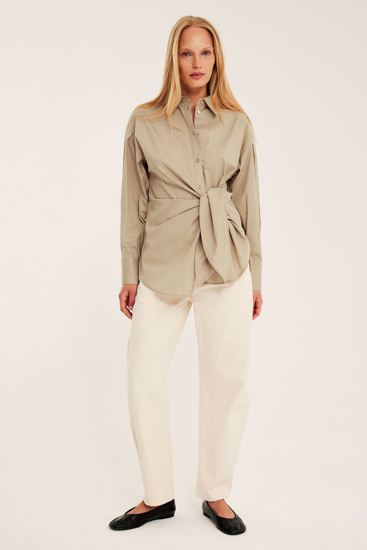 Edestä näkymä naisesta yllään beige puuvillapaita solmimalla edessä, kermanväriset leveälahkeiset housut ja mustat ballerinat.
