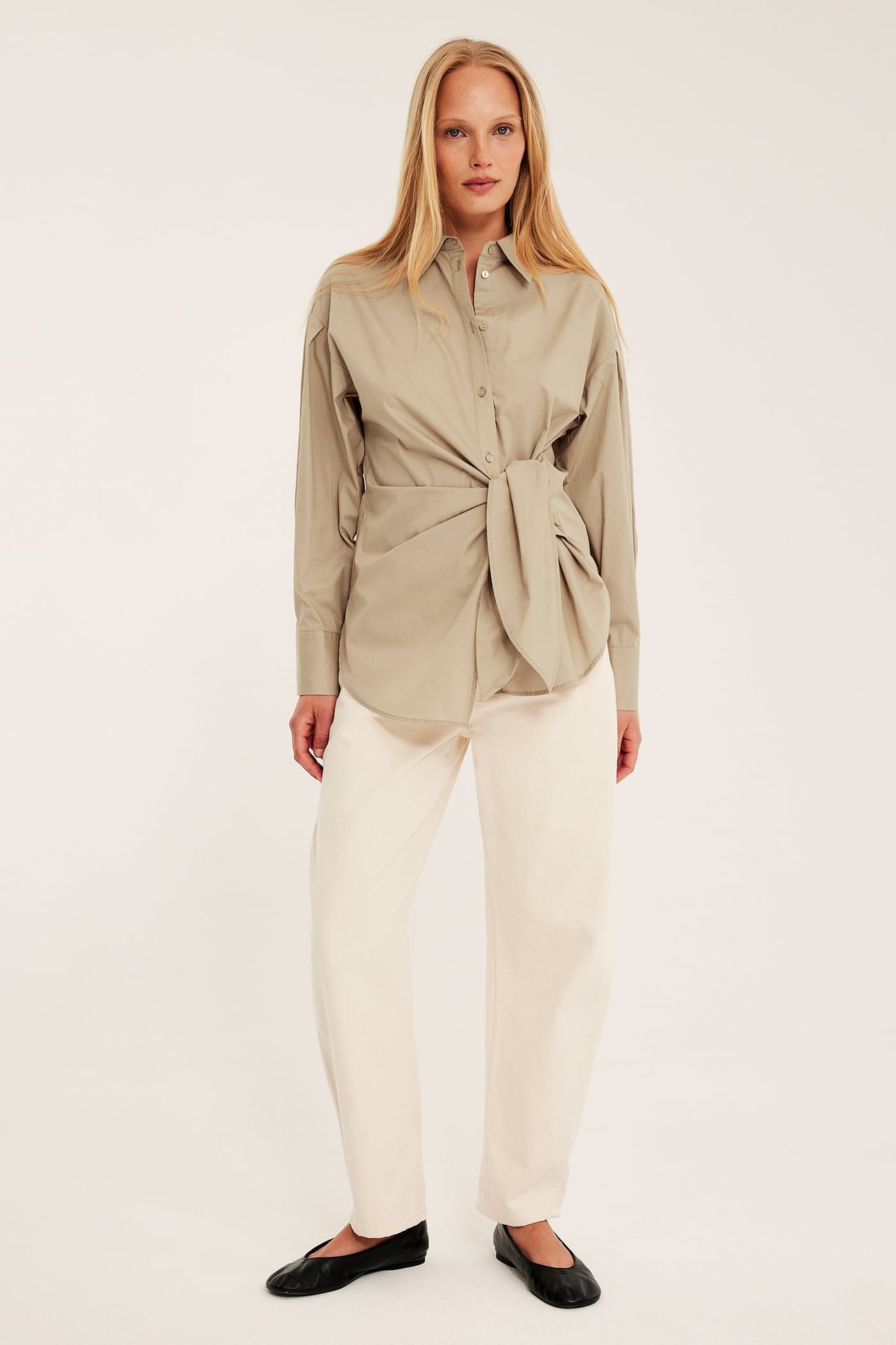 Edestä näkymä naisesta yllään beige puuvillapaita solmimalla edessä, kermanväriset leveälahkeiset housut ja mustat ballerinat.