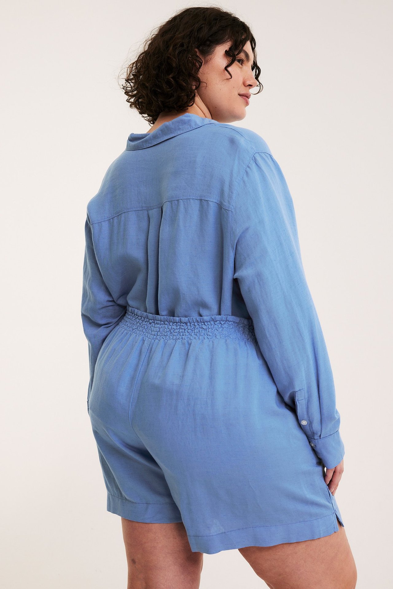 Vinklad ryggvy av en kvinna i blå viskos-linneshorts med smockresår och matchande långärmad skjorta.