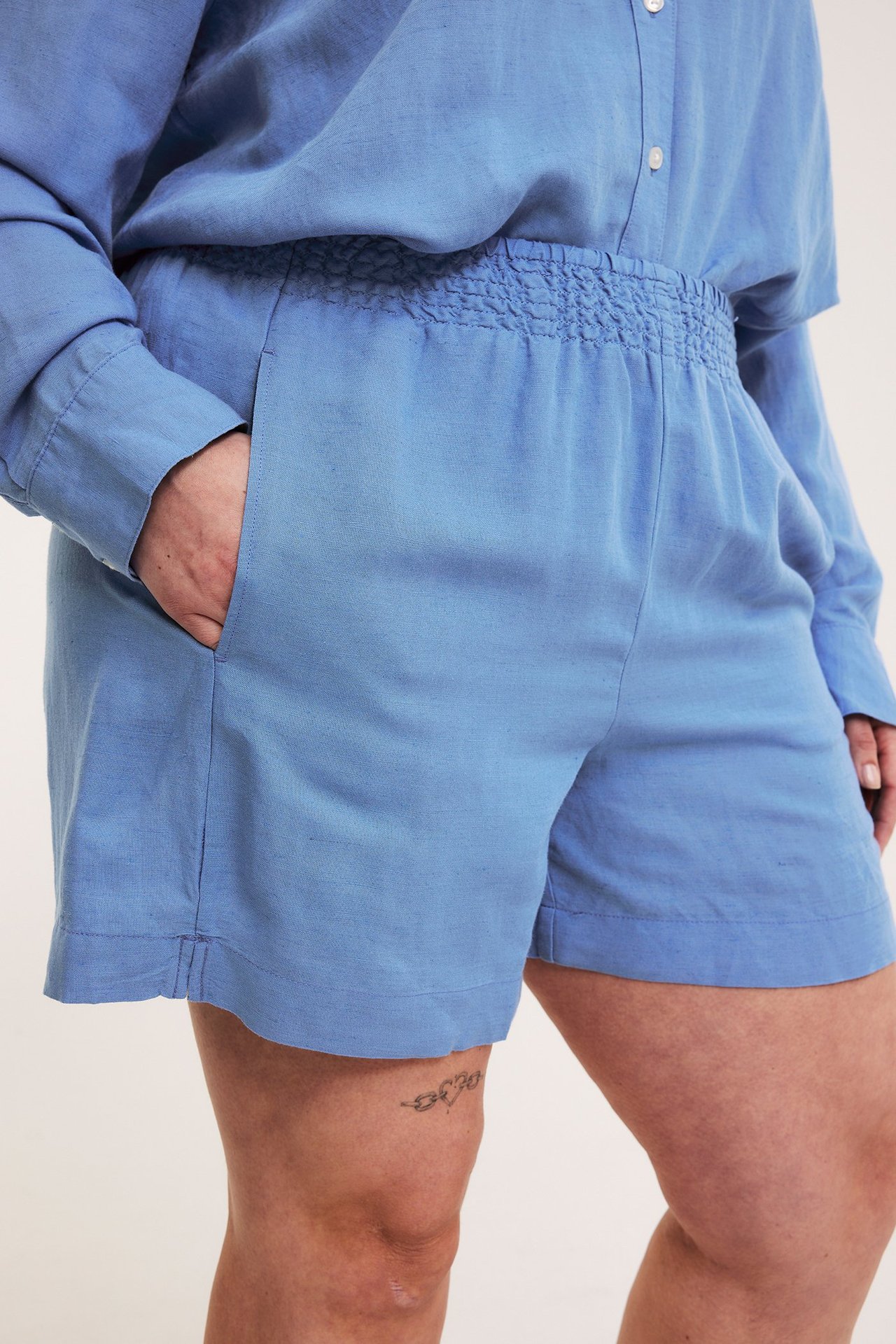 Närbild på kvinna i blå viskos-linneshorts med smockresår i midjan och matchande långärmad skjorta.