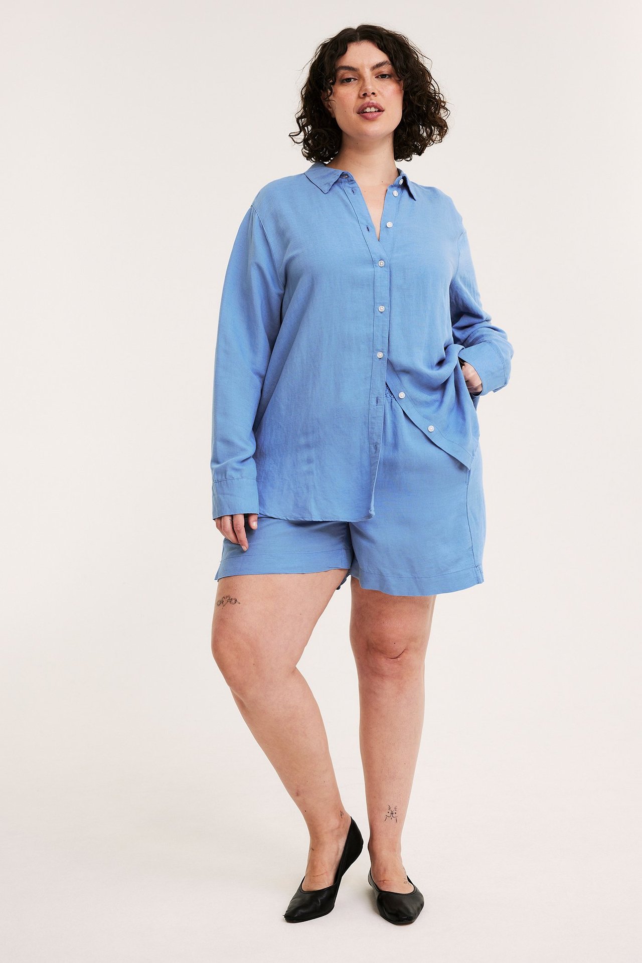 Forfra: kvinne i blå løse korte linblanding-shorts, matchende skjorte og svarte ballerinasko.
