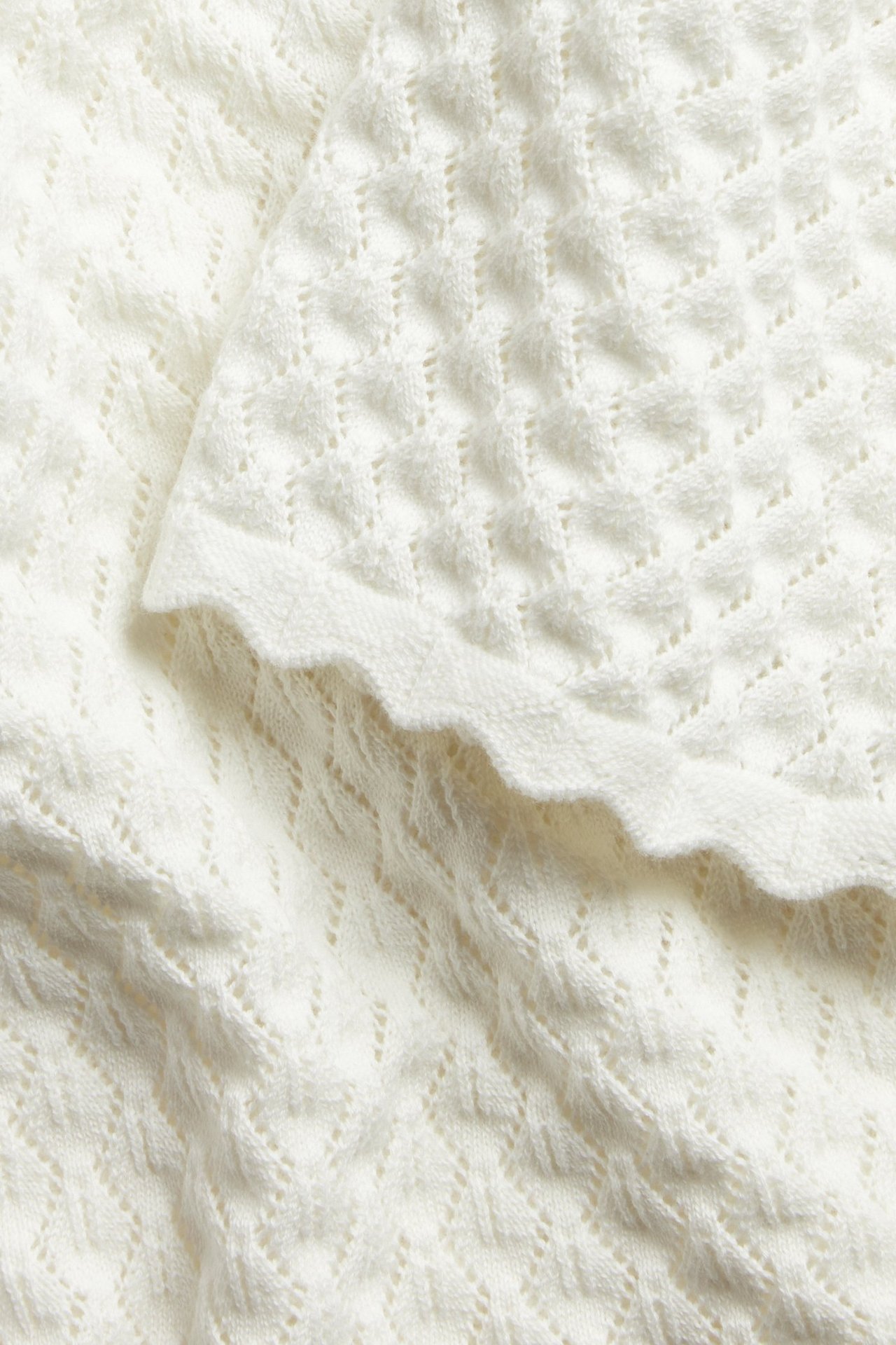 Zbliżenie na off-white dzianinowy materiał z krótkim rękawem, wyraźnym wzorem w romby i falistym brzegiem.