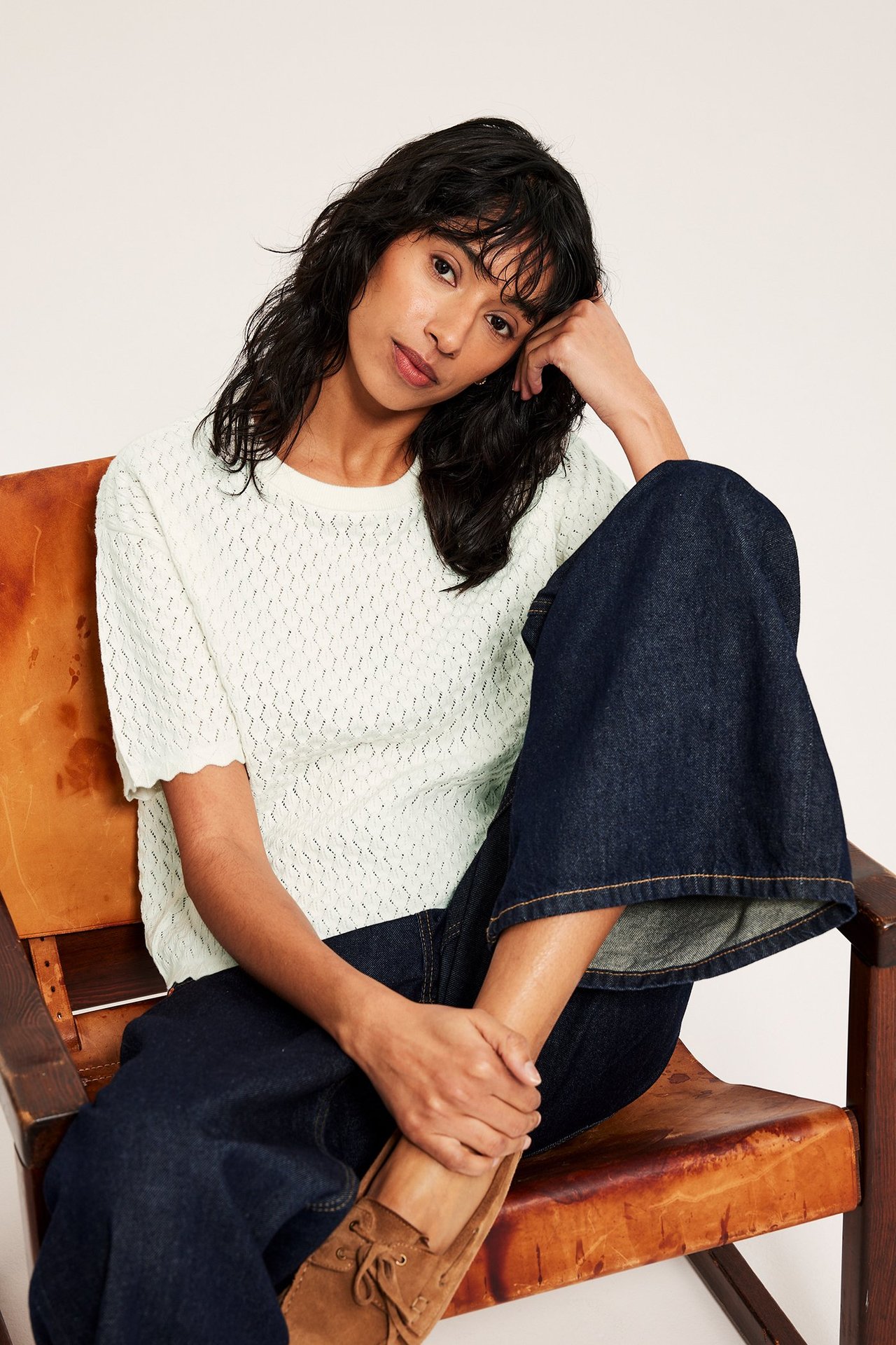 Frontvy av kvinna i offwhite mönsterstickad kortärmad tröja, mörkblå vida jeans och bruna mockaskor.