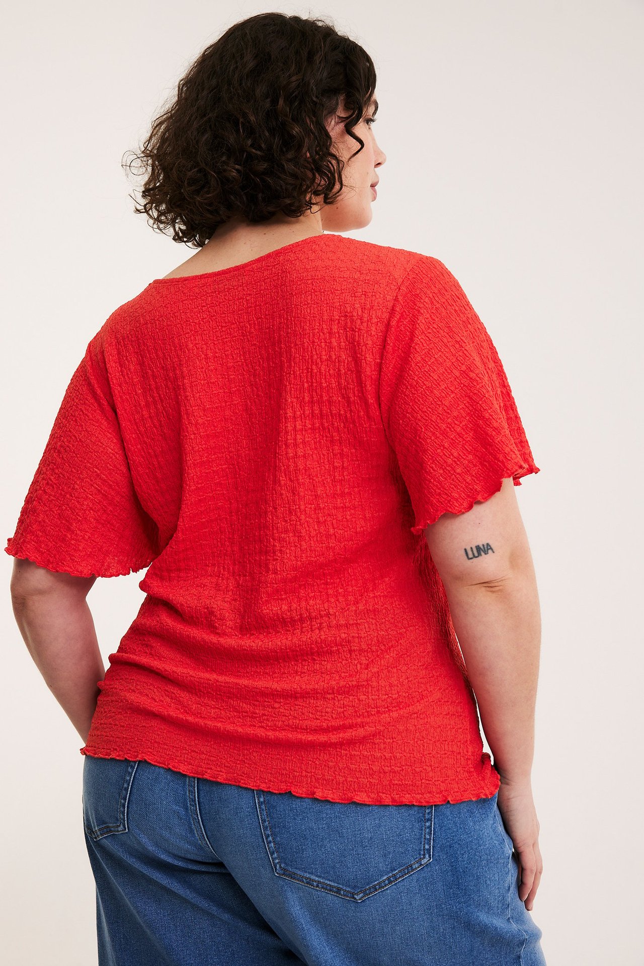 Naisen takakuva punaisessa strukturoidussa lyhythihaisessa topissa, jossa on aaltoilevat reunat, ja sinisissä farkuissa.