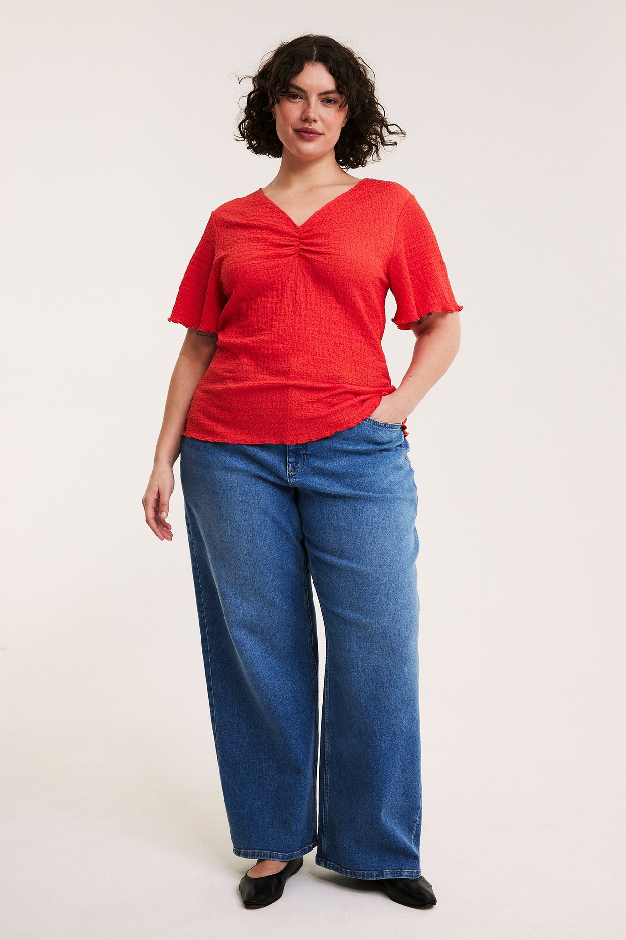 Edestä: Nainen punaisessa kuvioidussa V-kaula-aukkoisessa topissa, jossa frillahihat, sinisissä leveälahkeisissa farkuissa ja mustissa kengissä.