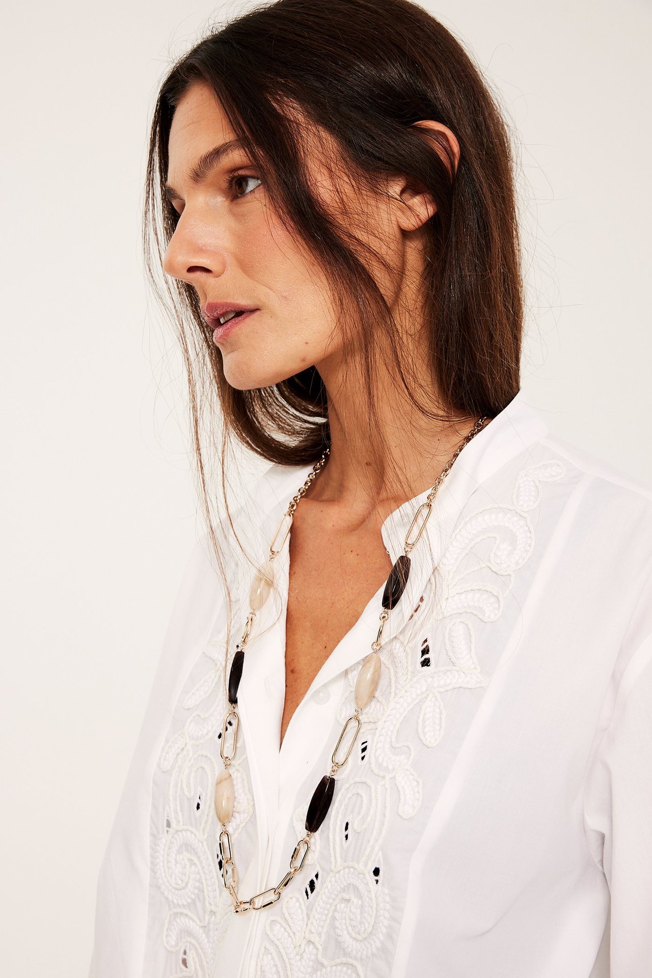 Lähikuva: Nainen kultakaulakoruineen, jossa vaaleat ja tummat soikeat kivet, valkoisen brodeeratun paidan päällä.