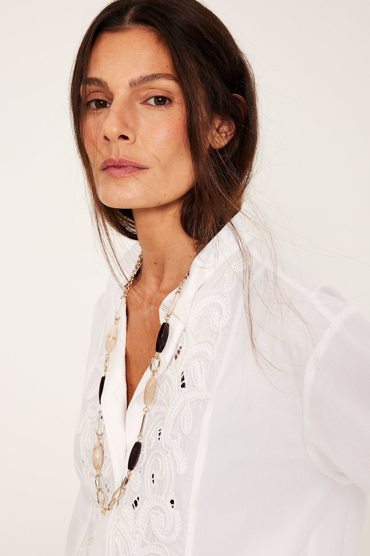 Lähikuva edestä: Nainen pitkä kultakaulakoru vaaleilla ja tummilla kivillä valkoisen brodeeratun puseron päällä.