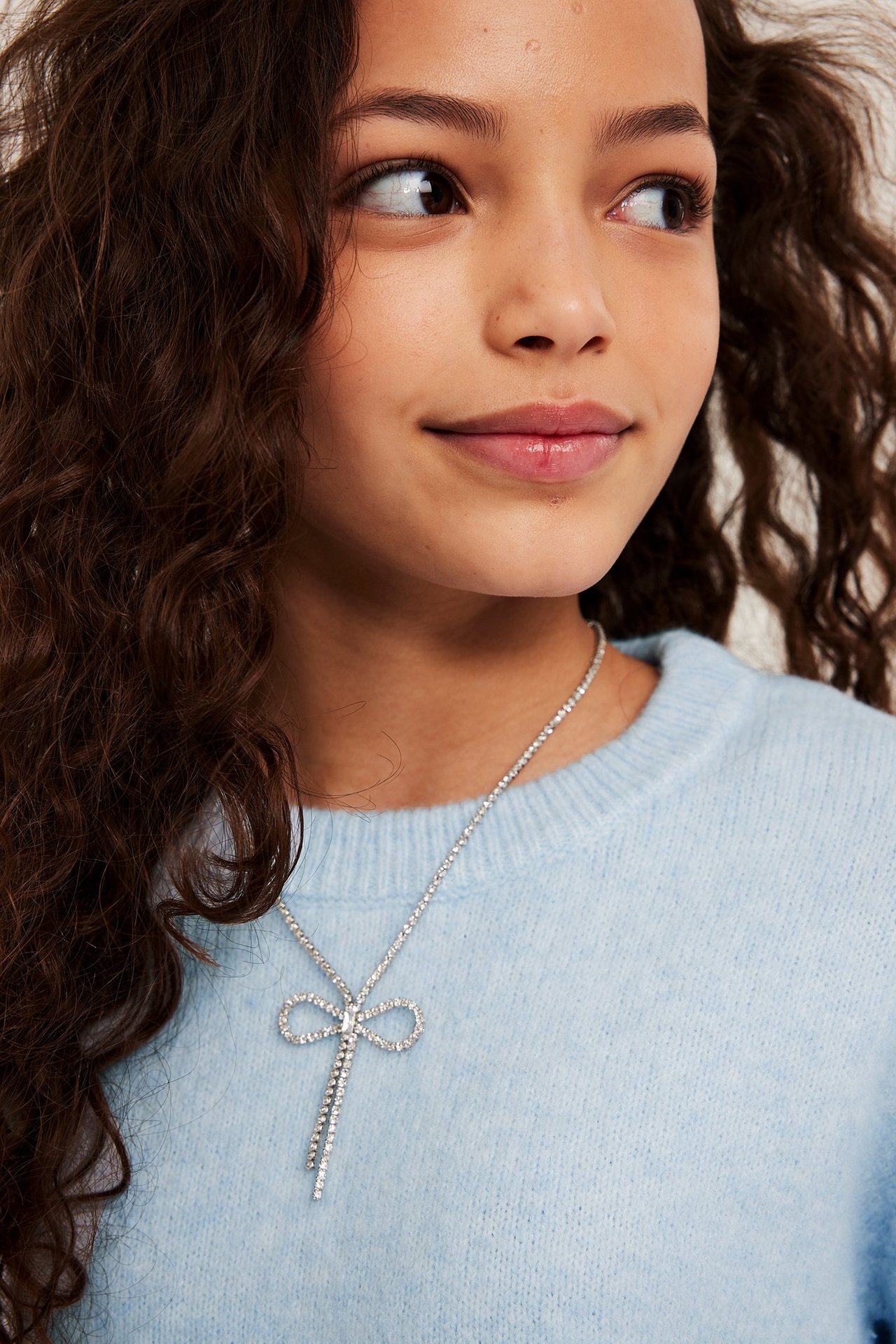 Nærbilde av et barn med et sølvfarget strassløyfehalskjede over en lyseblå finstrikket genser.