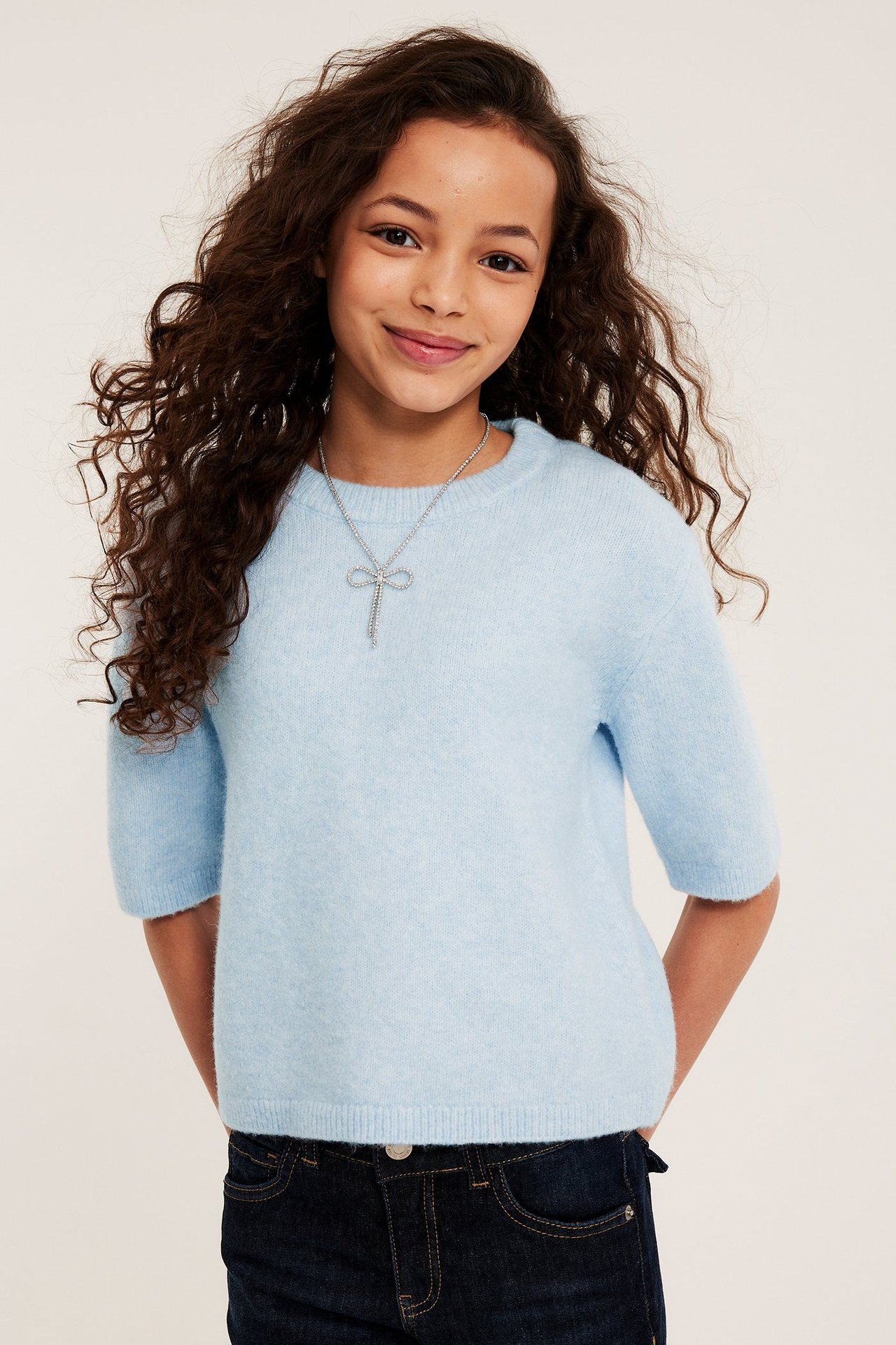 Widok z przodu dziecka w jasnoniebieskim swetrze z krótkim rękawem, ciemnych dżinsach i srebrnym naszyjniku kokardce.
