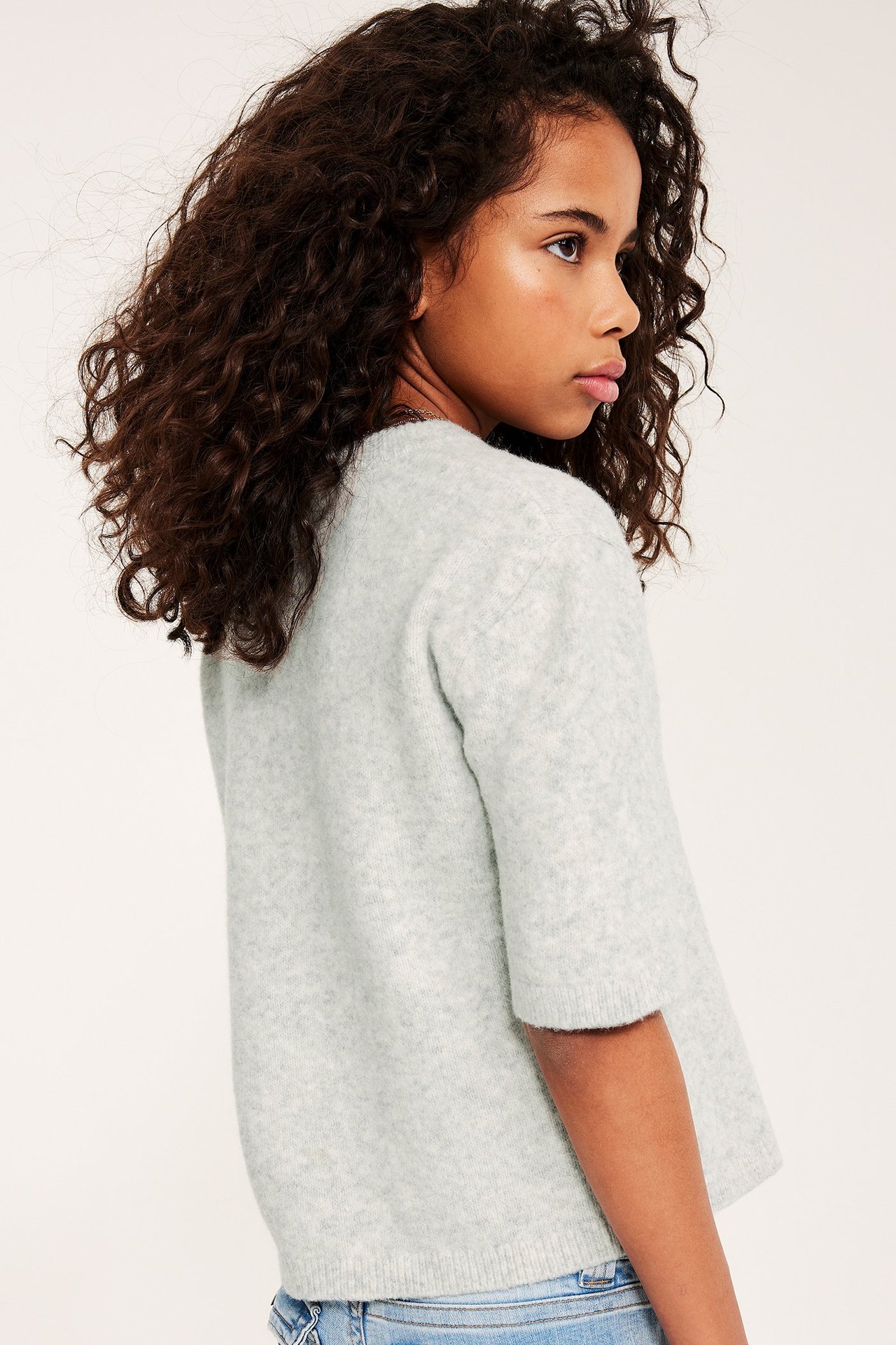 Widok z boku dziecka w jasnoszarym melanżowym, cienkim swetrze z krótkim rękawem, na jasnych dżinsach.