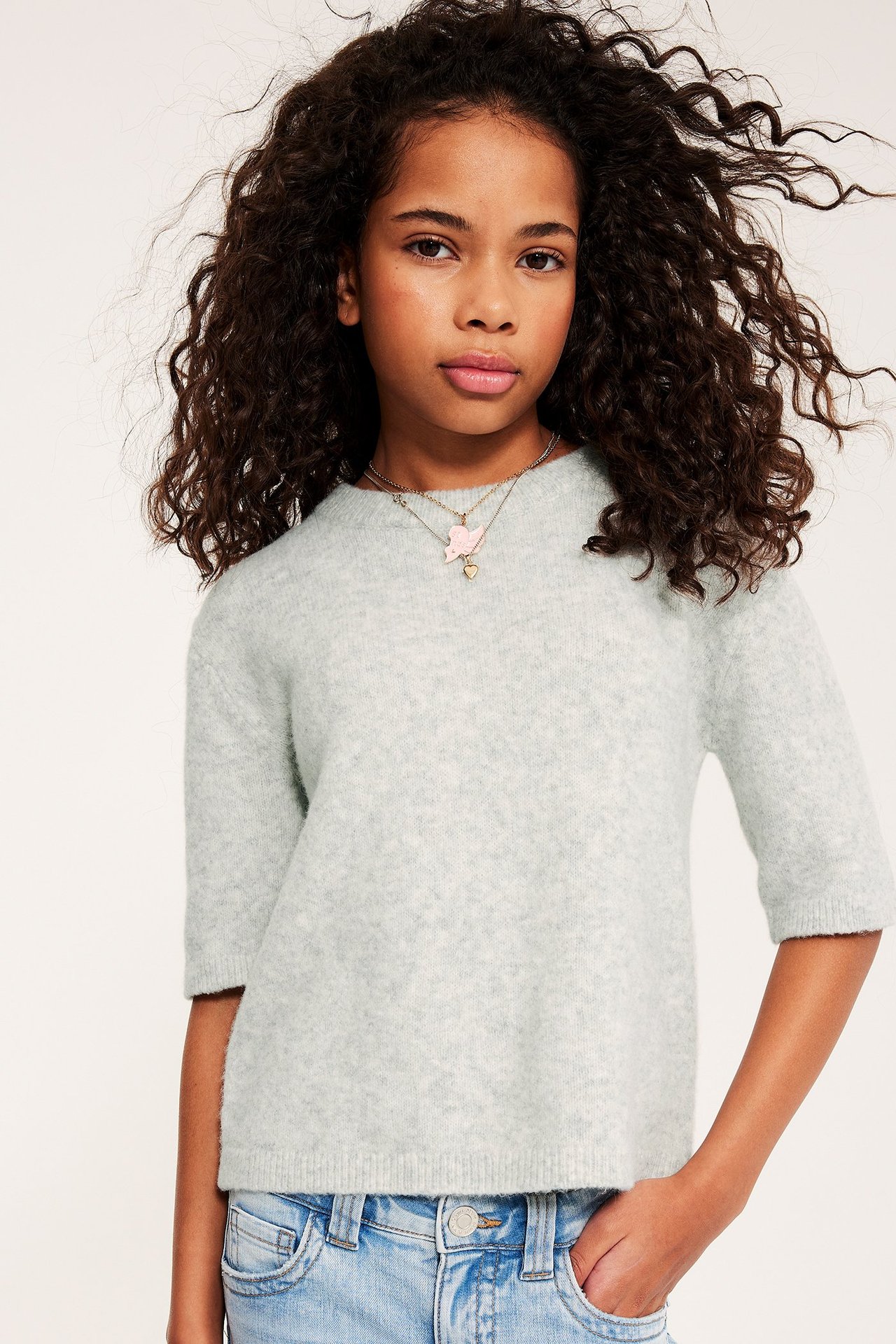 Frontbilde av et barn i en lys grå melert kortermet genser, lyseblå jeans og halskjeder.