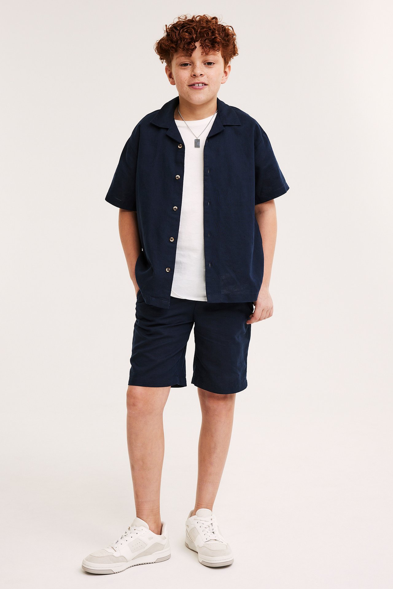 Pojke bär mörkblå shorts i linneblandning, matchande öppen skjorta, vit t-shirt, vita sneakers och halsband.