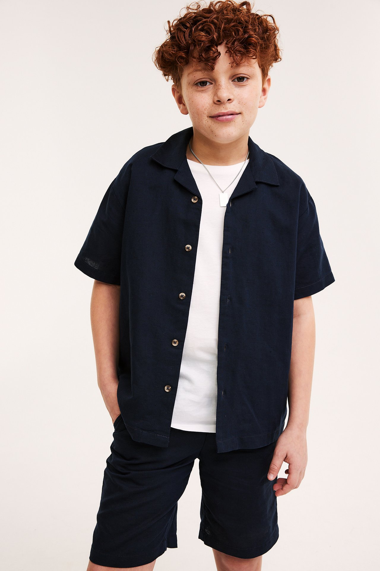 Framifrån, ett barn i mörkblå shorts i linnemix med resårmidja, en matchande öppen skjorta och vit t-shirt.