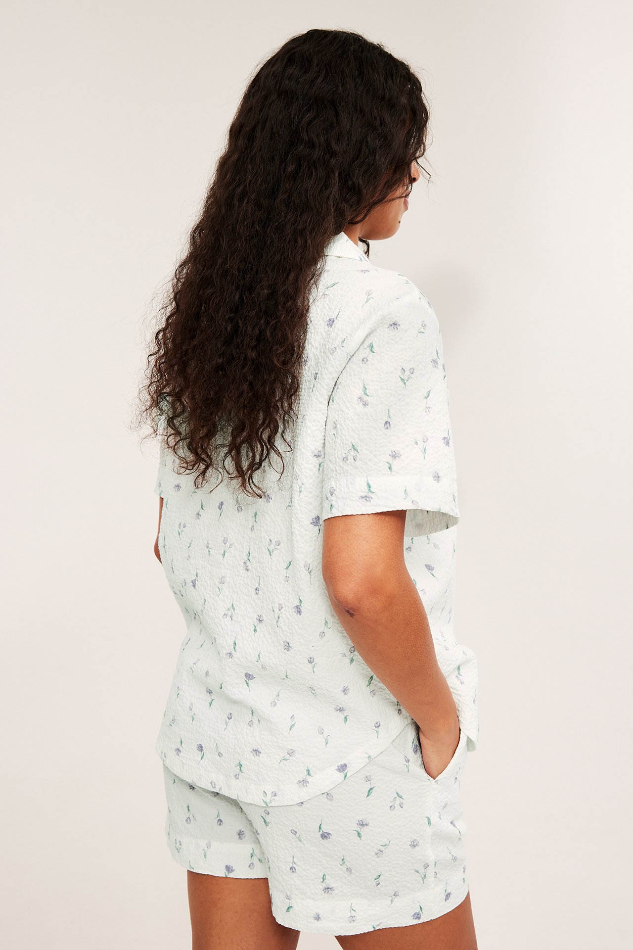 Sidovy av kvinna i offwhite blommigt, strukturerat kortärmat pyjamasset, med handen i fickan.