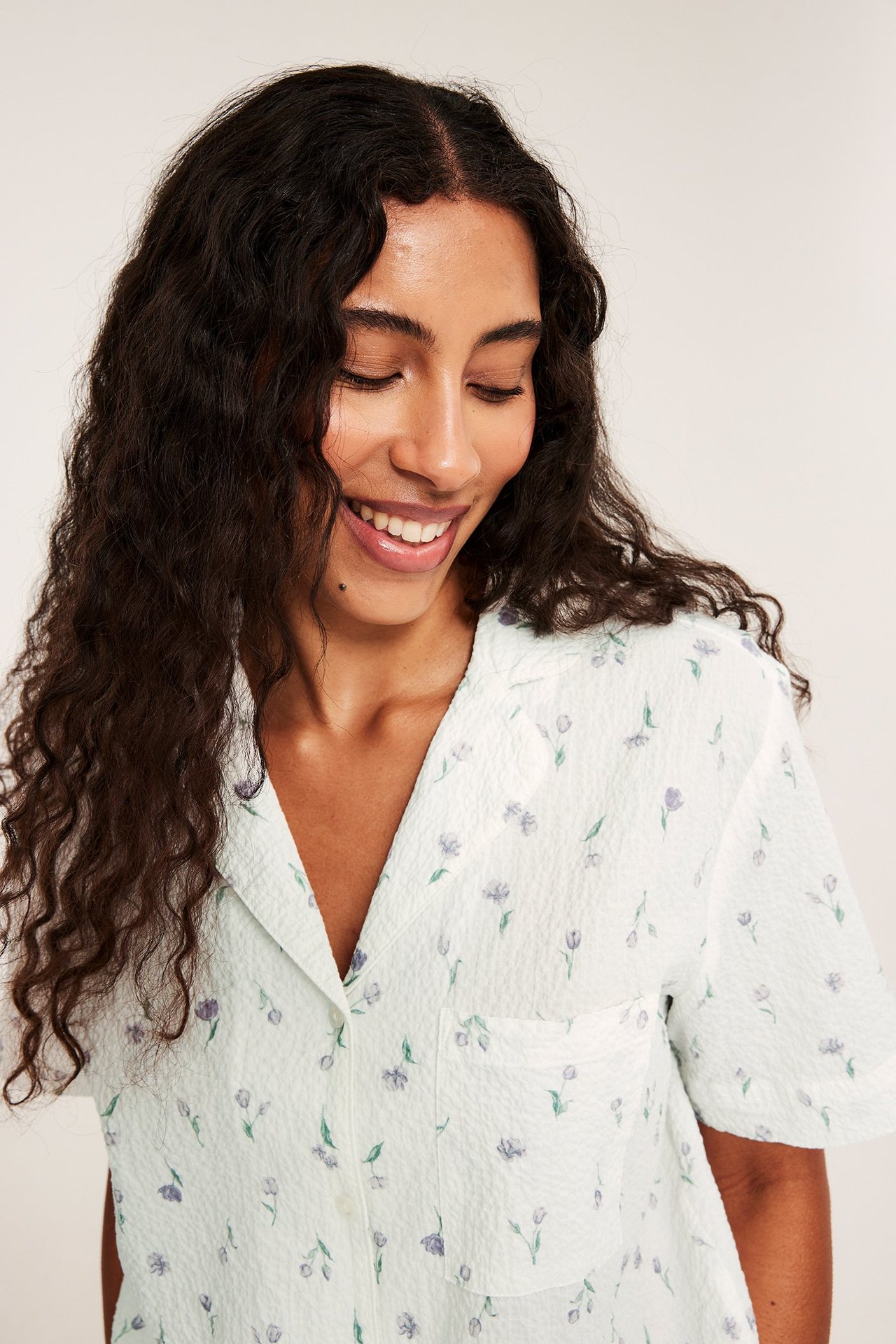 Forfra: kvinne i en offwhite krøllete kortermet pyjamasoverdel med lilla blomstermønster.