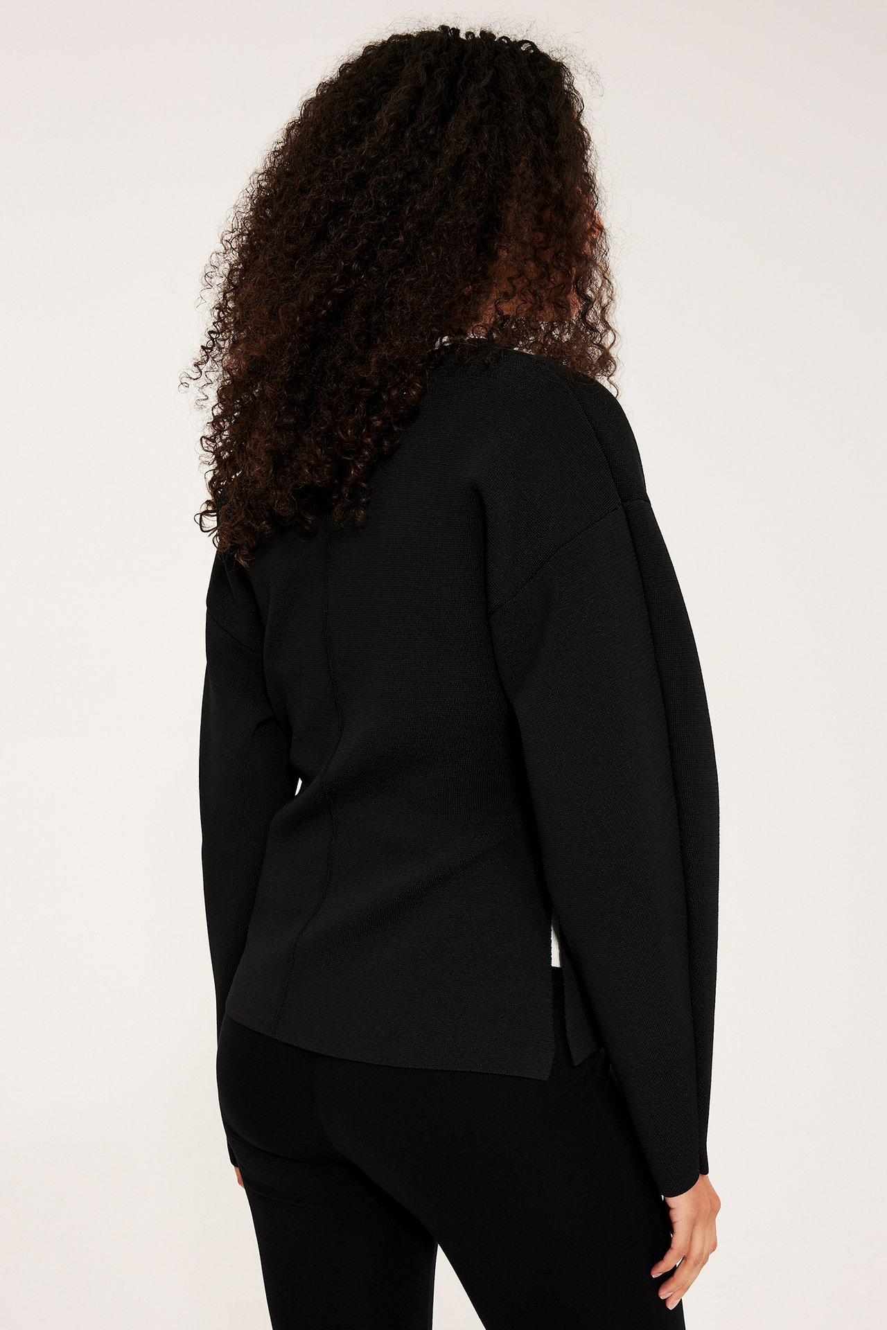 Ryggvy av kvinna i svart långärmad kofta med formad midja över svarta byxor.
