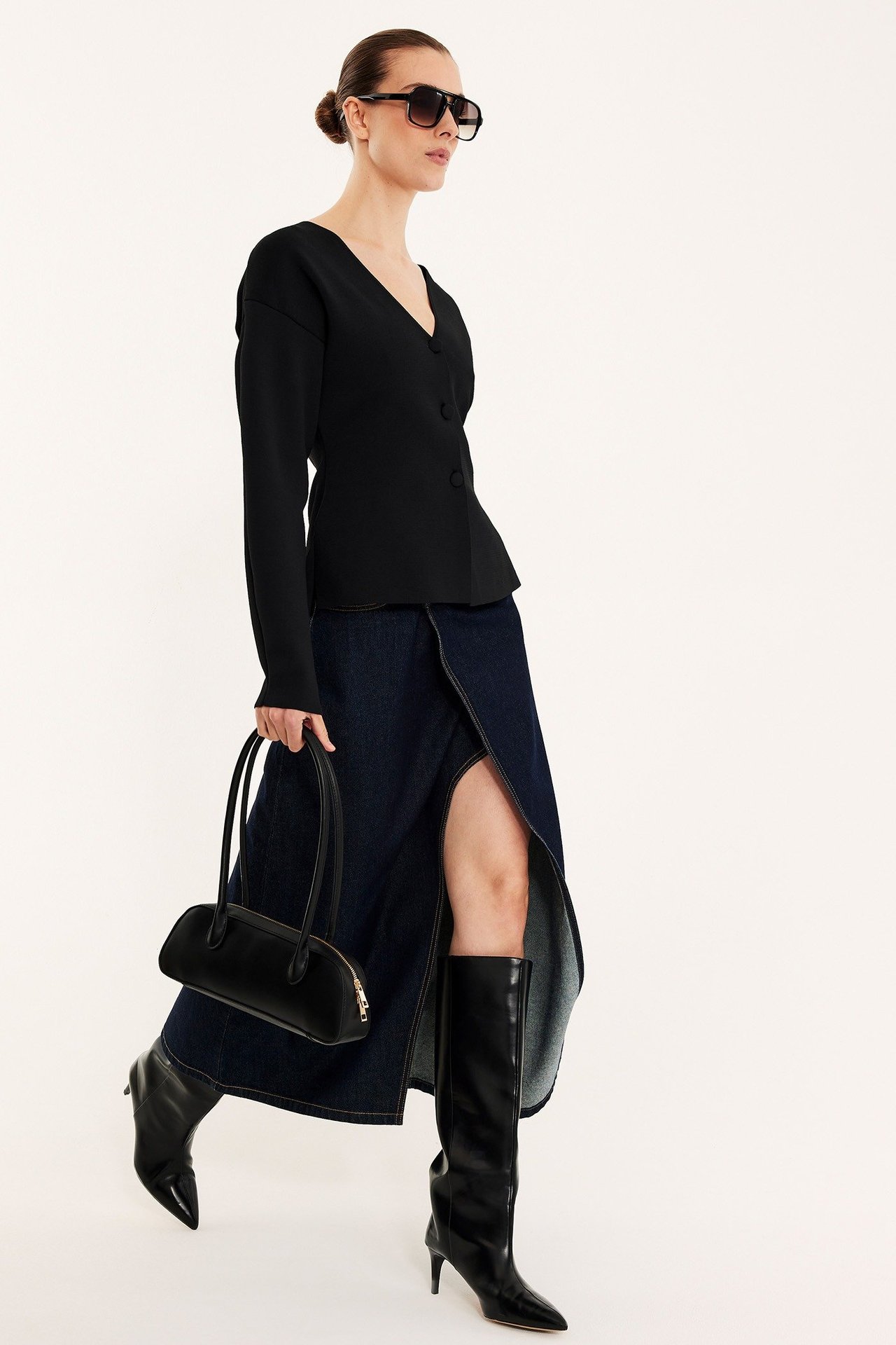 Nainen mustassa muotoonleikatussa neuletakissa, tummassa farkkuhameessa, mustissa polvisaappaissa ja mustassa olkalaukussa.