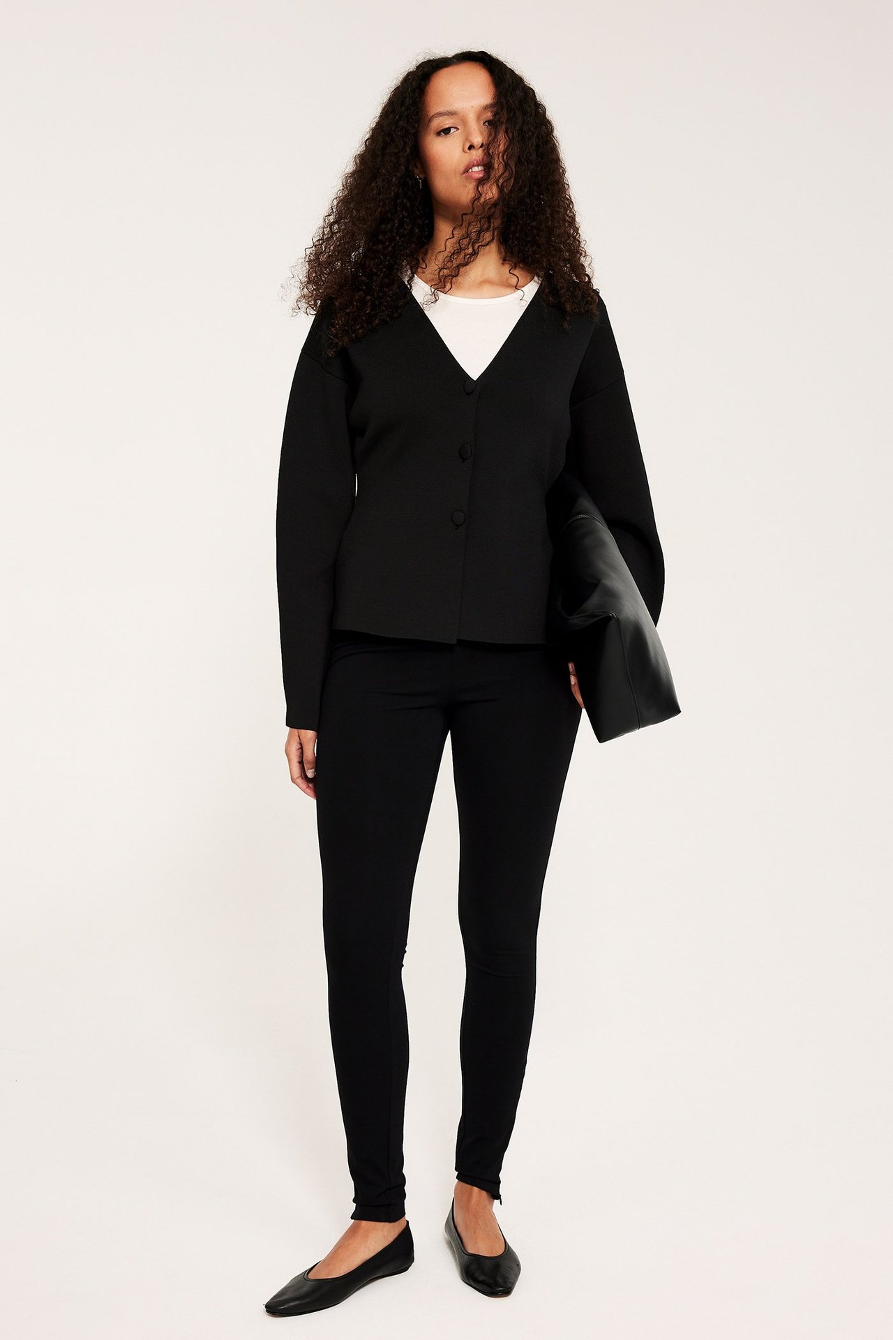 Kvinne i svart formet cardigan, hvit topp, svarte smale bukser, svarte ballerinasko, holder svart clutch.