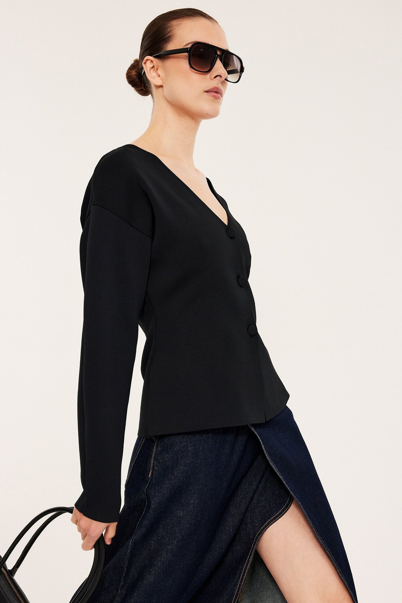 Sidebilde av en kvinne i svart formsydd cardigan med knapper, mørkt dongeriskjørt og svarte solbriller.