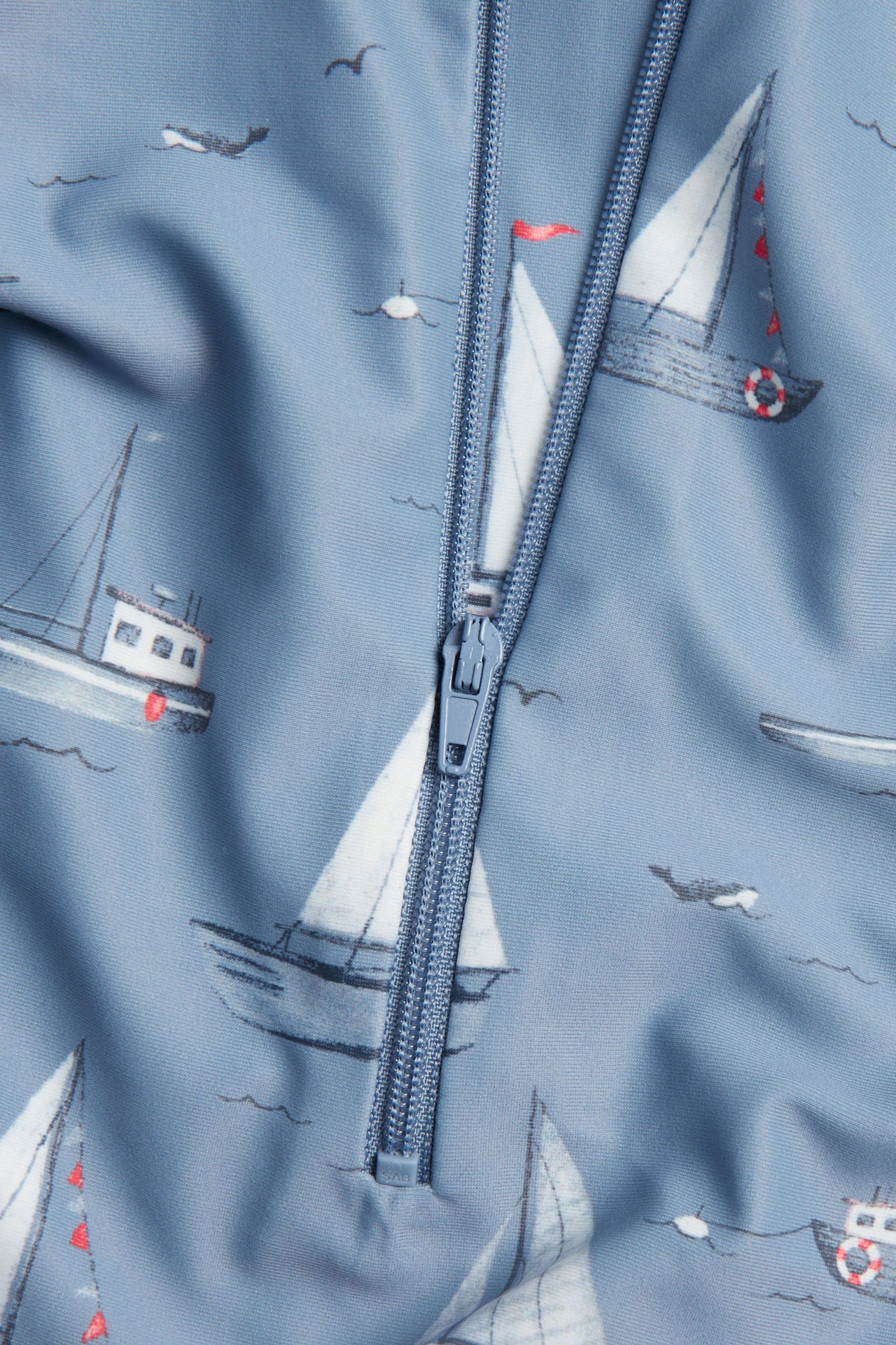 Närbild av en blå baby/barn UV-dräkt med vitt båtmönster och synlig blå dragkedja.