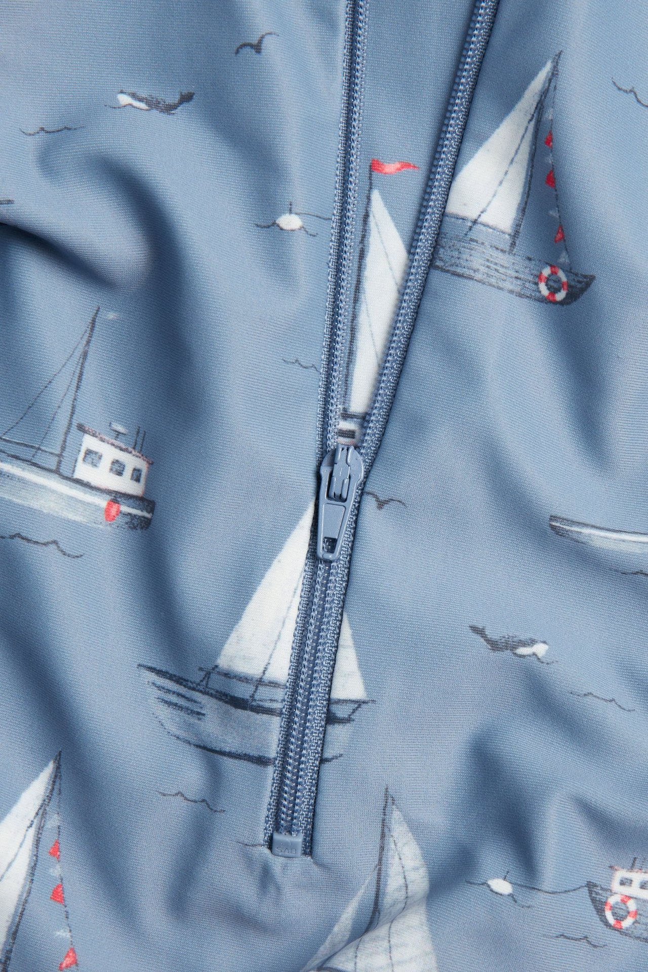 Nærbilde av en blå baby/barn UV-romper med hvitt båtmønster og synlig blå glidelås.