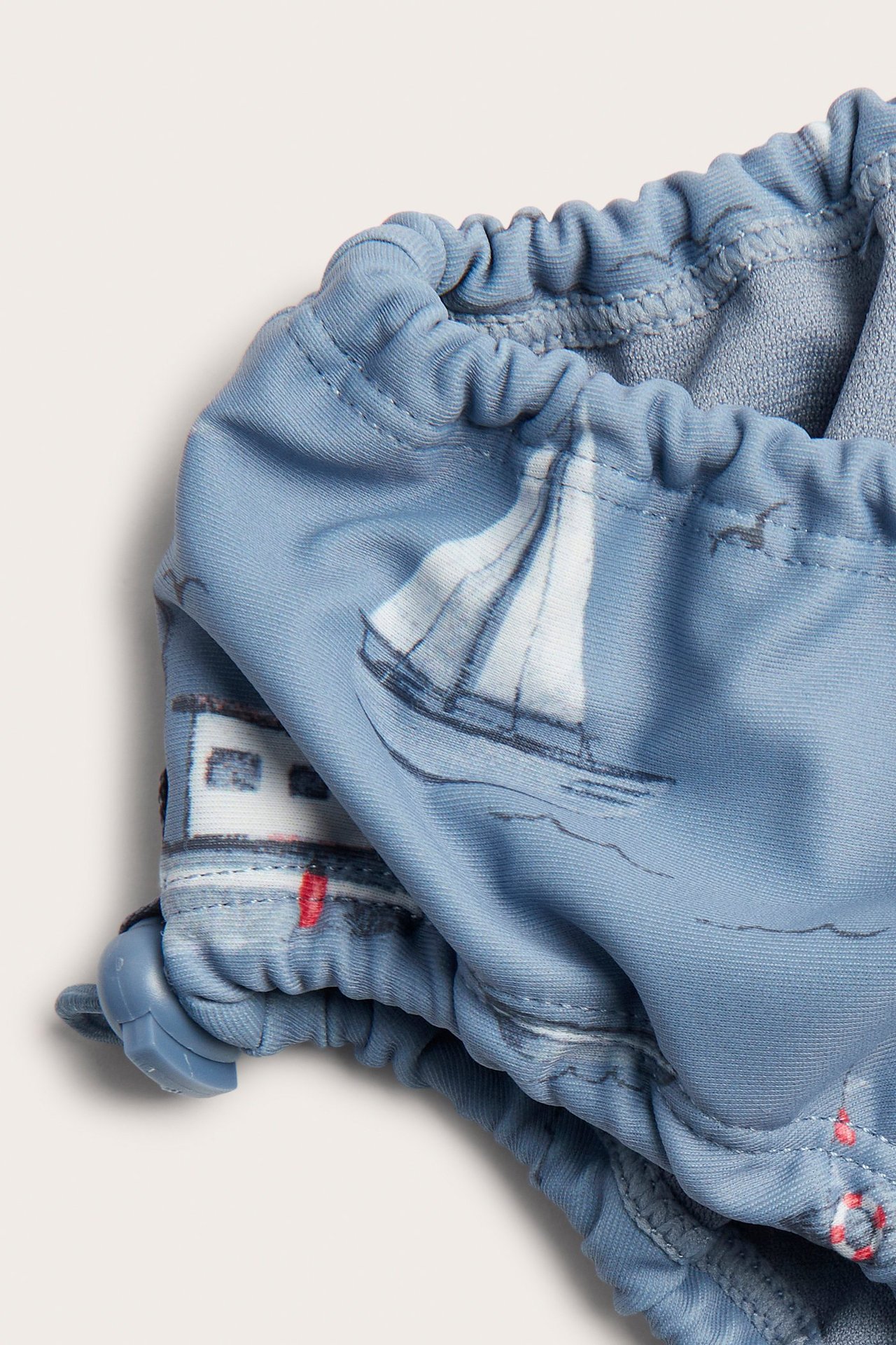 Nærbilde av blå babybadebukser med båtmønster, elastiske kanter og justerbare stoppere.