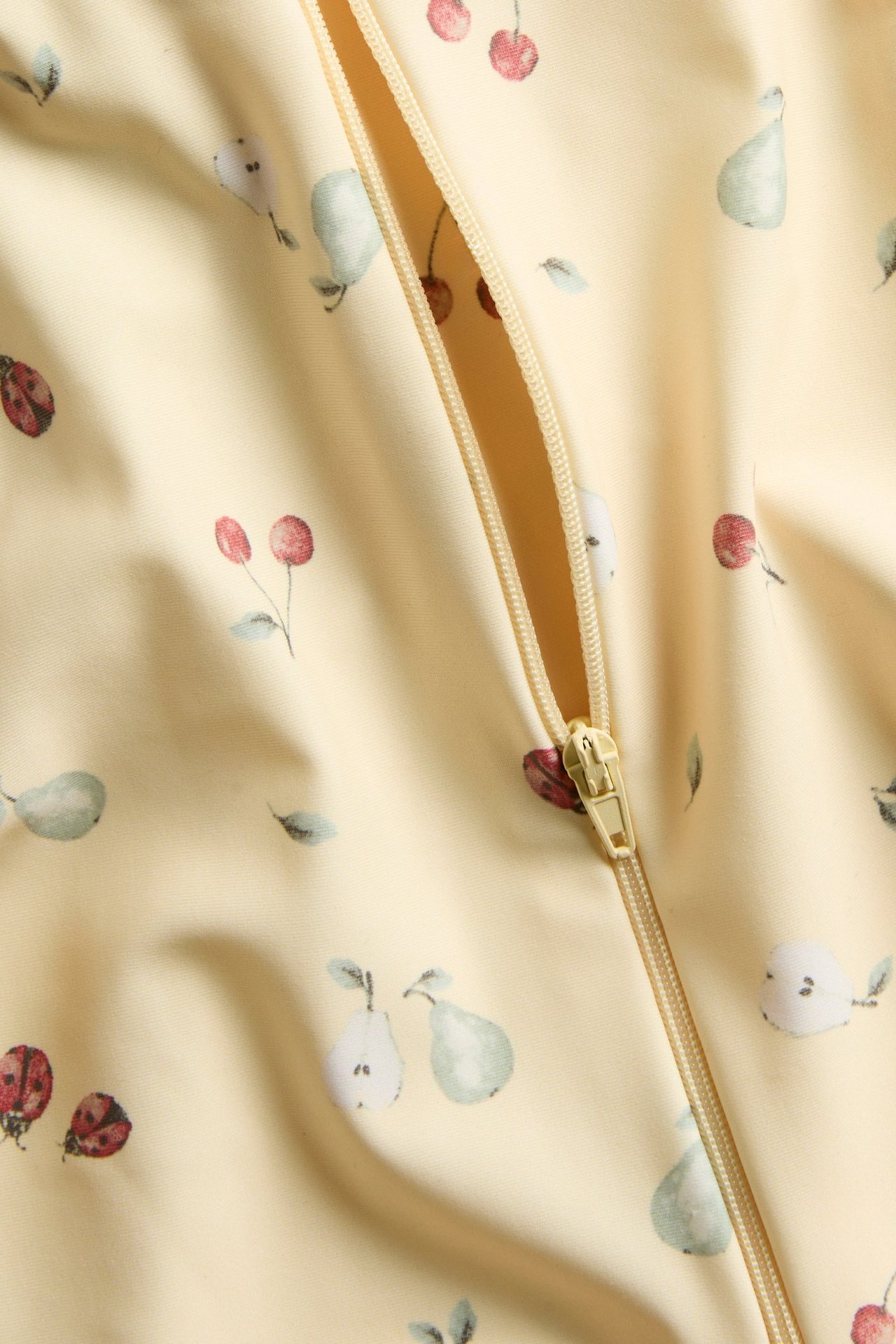 Närbild på en gul mönstrad UV-dräkt med körsbär, päron och nyckelpigor, med dragkedja.