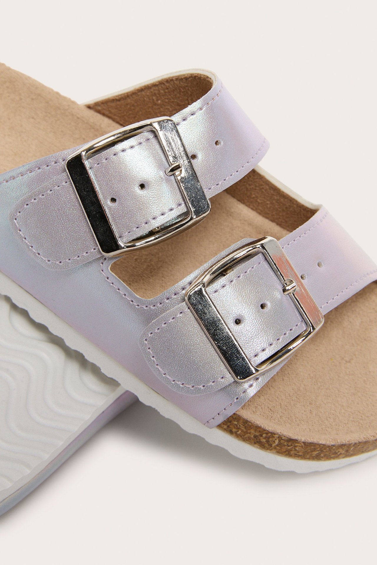 Zbliżenie na mieniące się sandały z dwiema regulowanymi srebrnymi klamrami i korkową podeszwą.