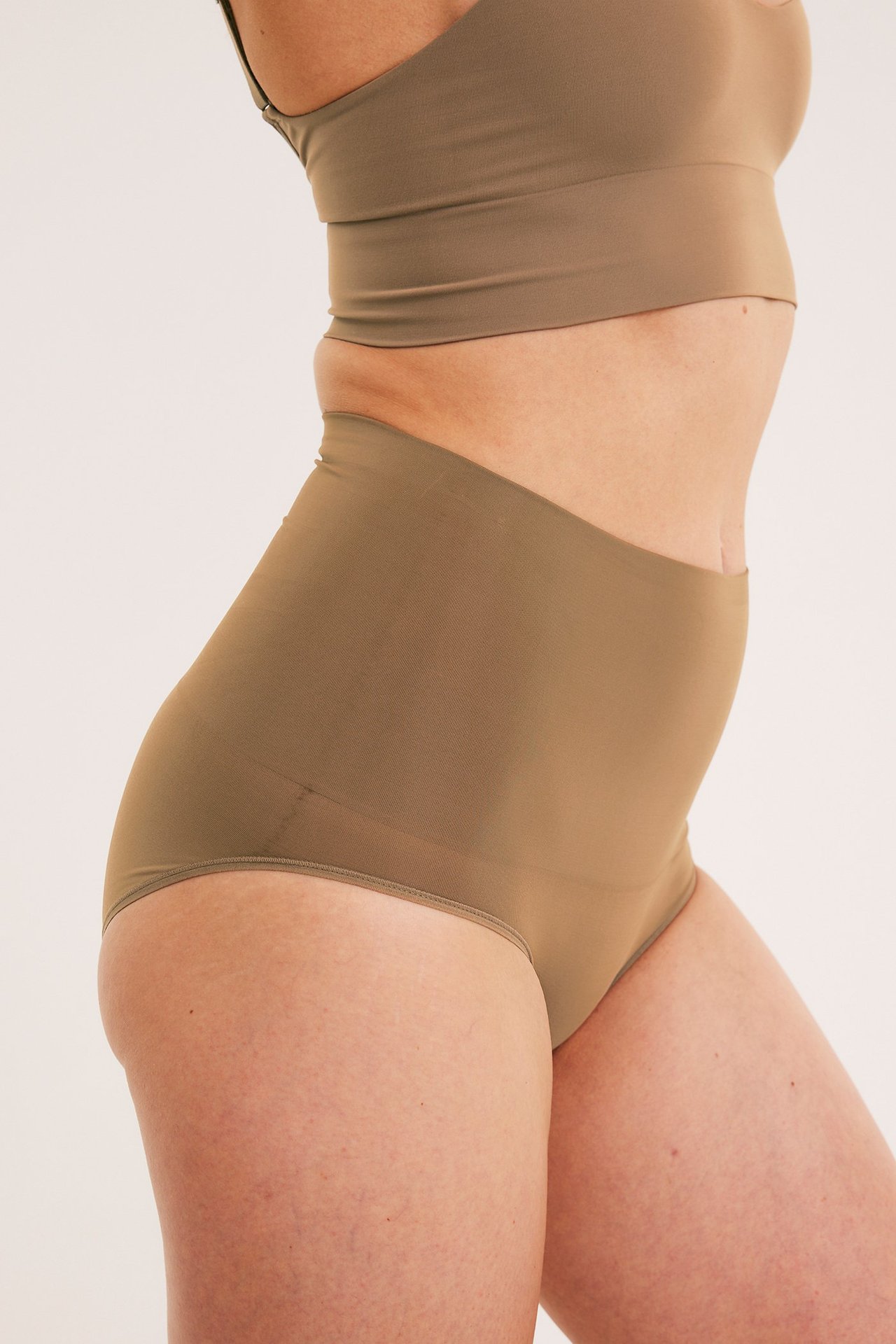 Sivukuva naisesta yllään korkeavyötäröiset ruskeat muotoilevat alushousut ja yhteensopiva crop top.