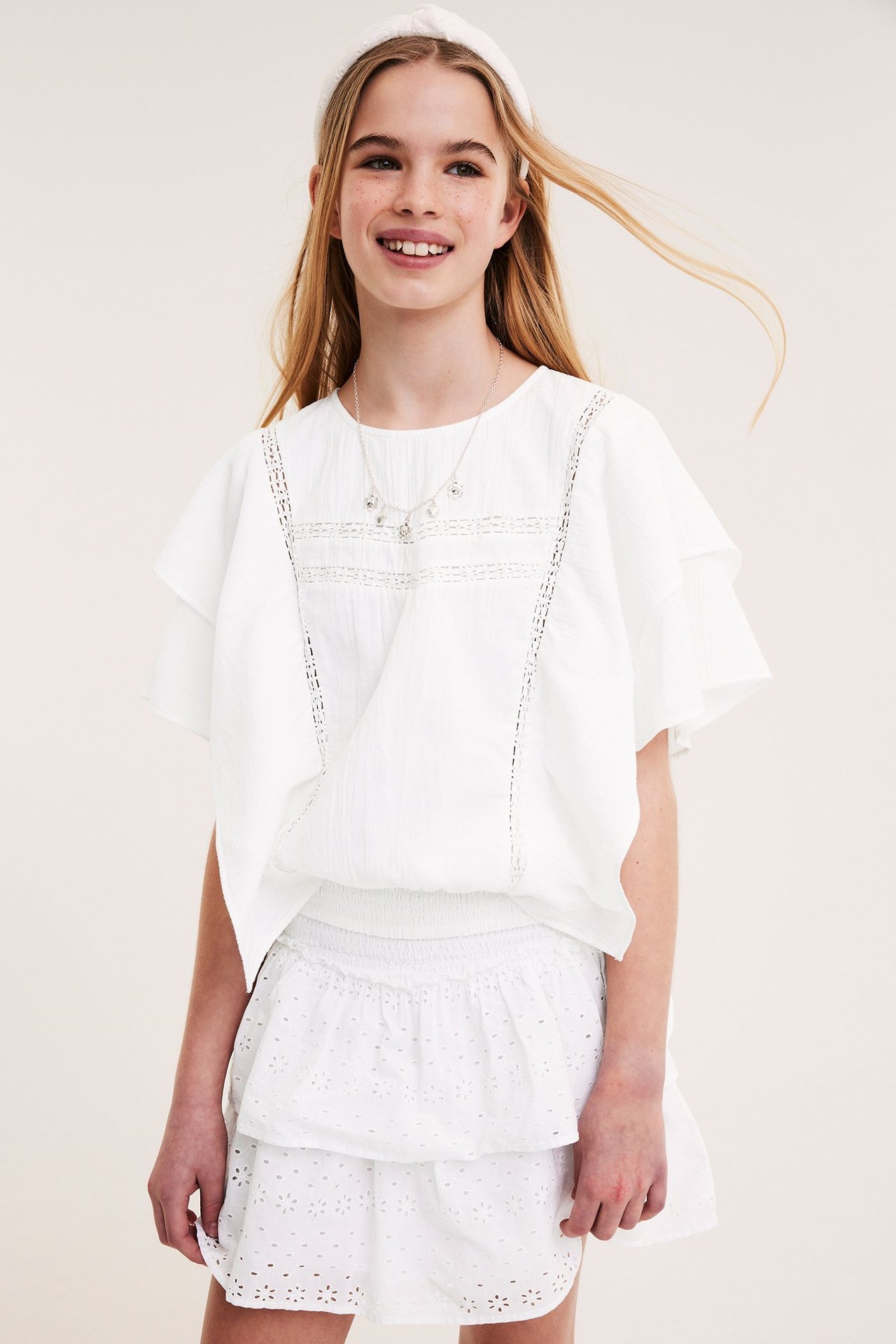 Widok z przodu: Dziecko nosi białą bawełnianą bluzkę z koronką i marszczeniem, białą spódnicę, naszyjnik, opaskę.