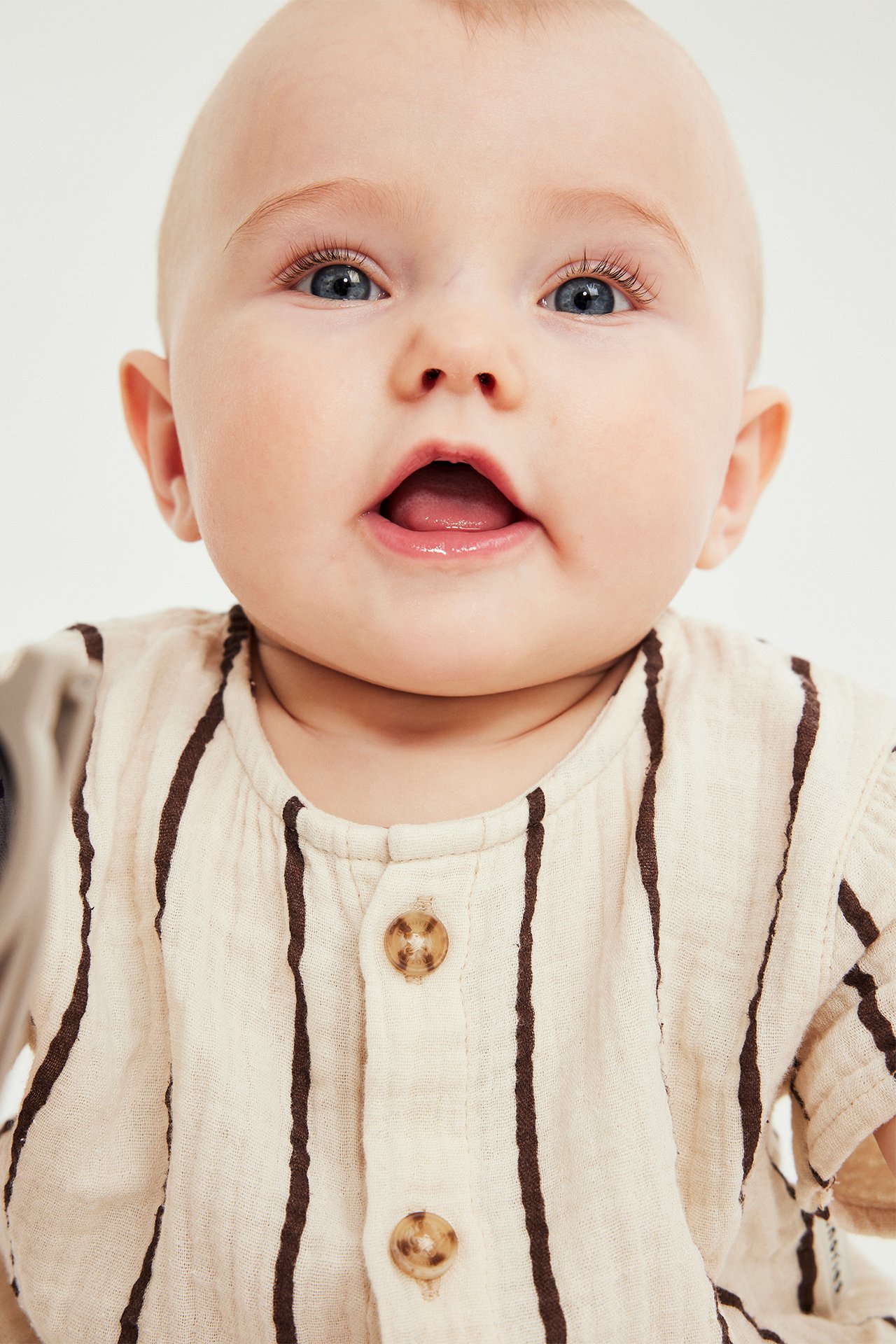 Nærbilde forfra av baby i stripete beige kortermet musselinromper med brune knapper.