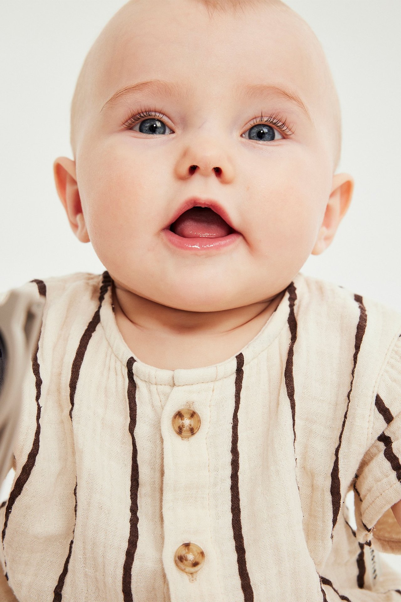 Närbild framifrån på en baby i randig beige kortärmad muslinromper med bruna knappar.