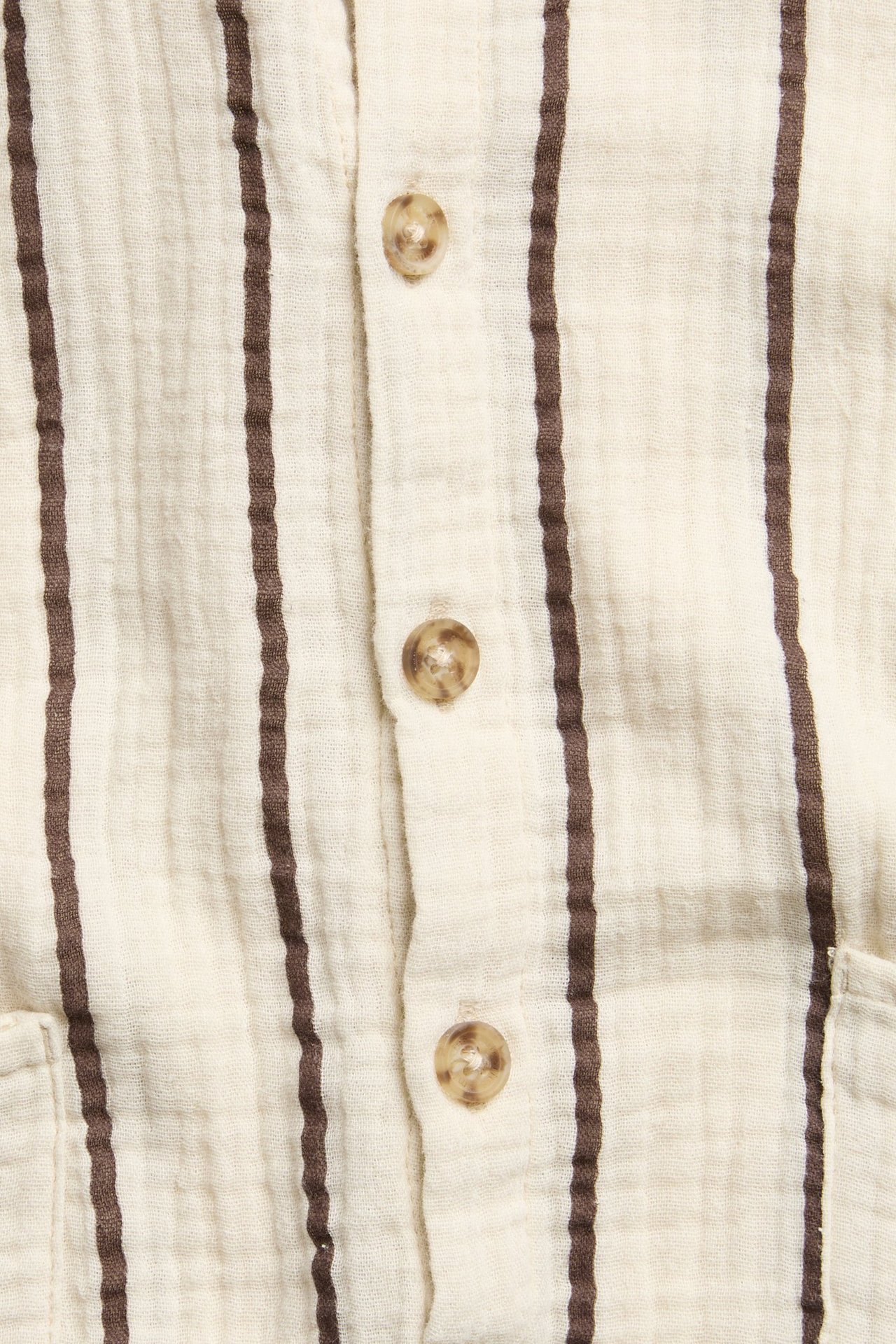 Lähikuva: beige raidallinen musliini-vauvanhaalari edessä napeilla ja näkyvällä taskulla.