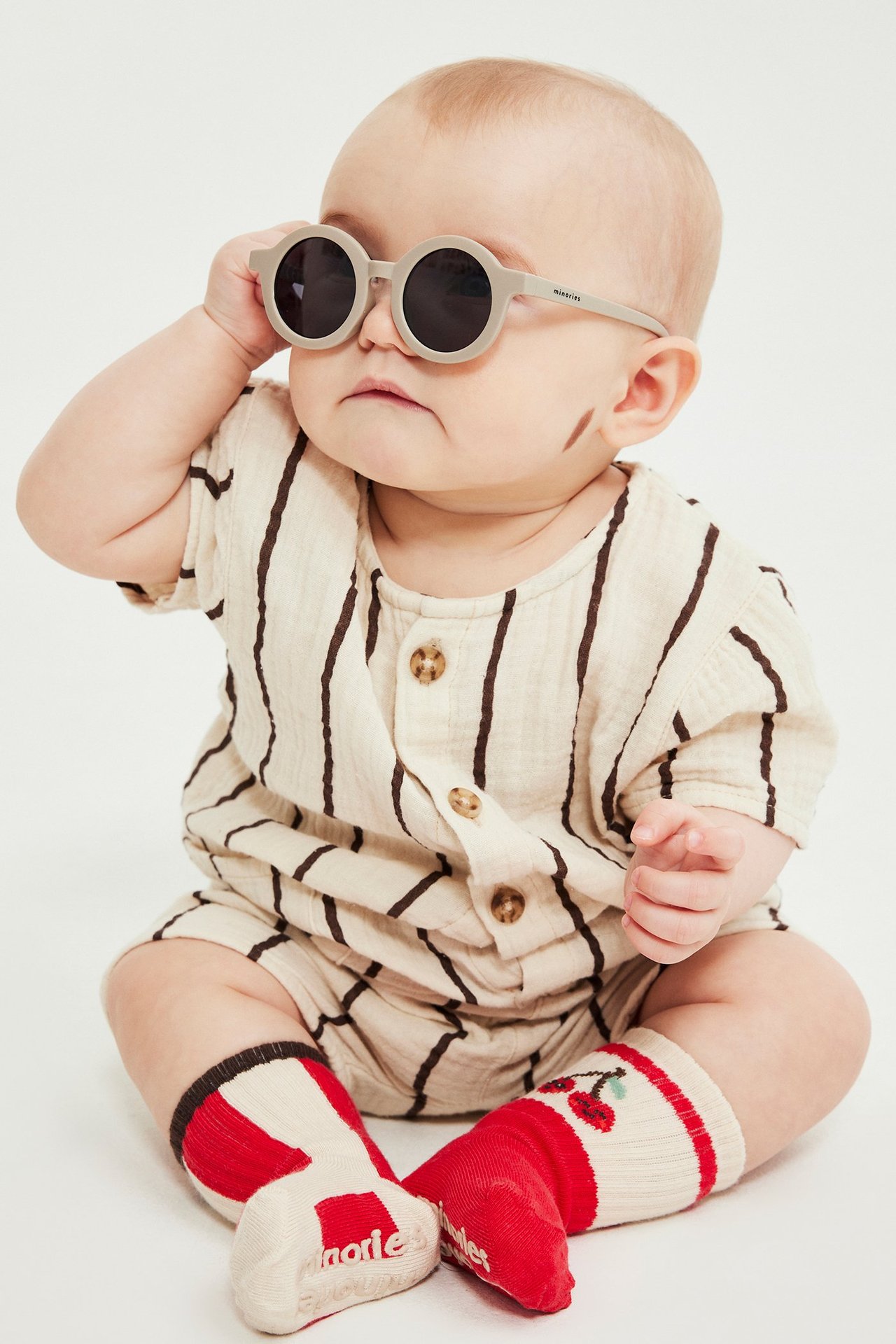 Frontvy av baby i randig beige muslin-romper med knappar, grå runda solglasögon och röda körsbärsstrumpor.