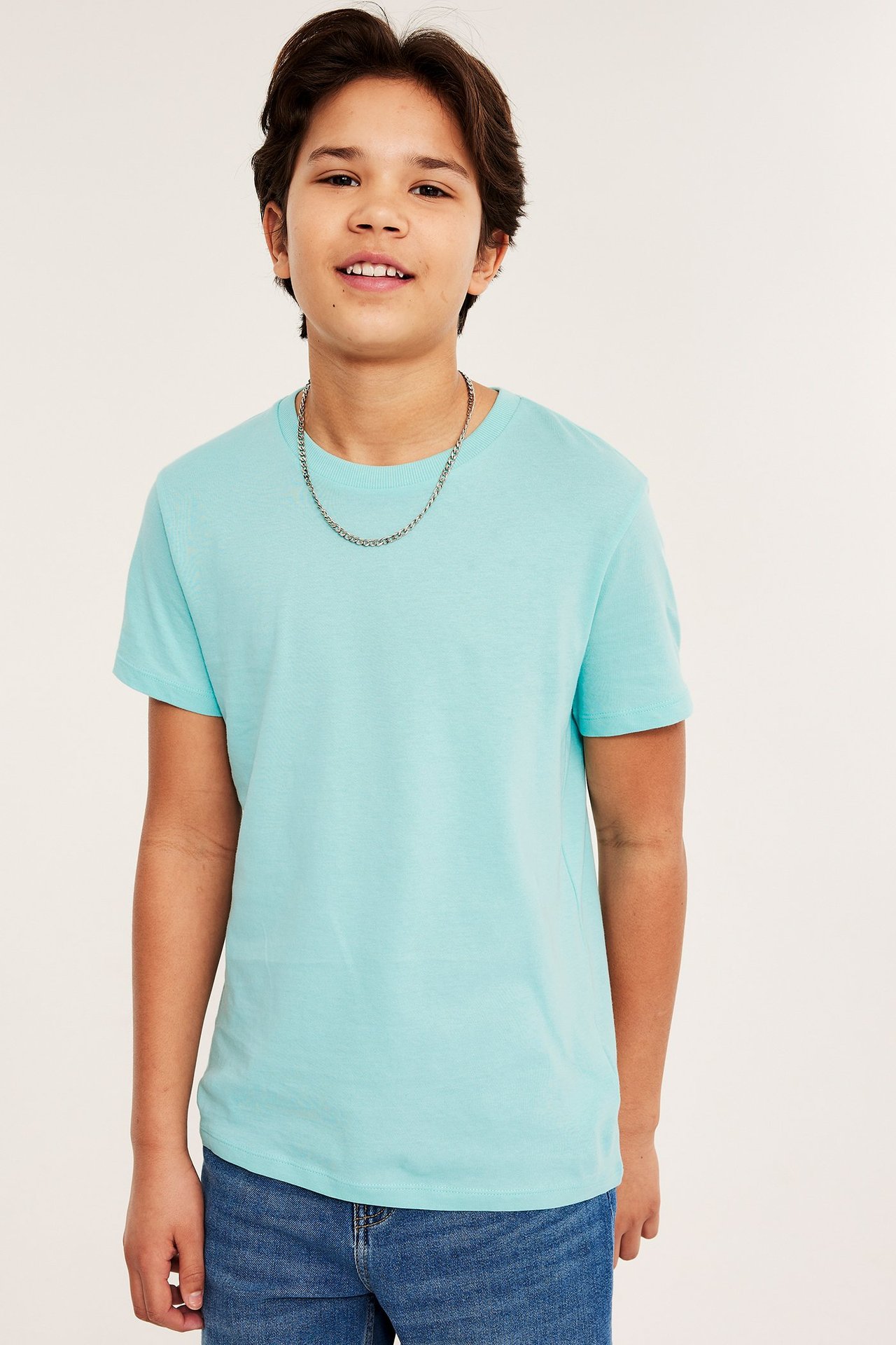 Edestä: Lapsi yllään vaaleanturkoosi puuvillainen t-paita, hopeanvärinen ketju ja siniset farkut.