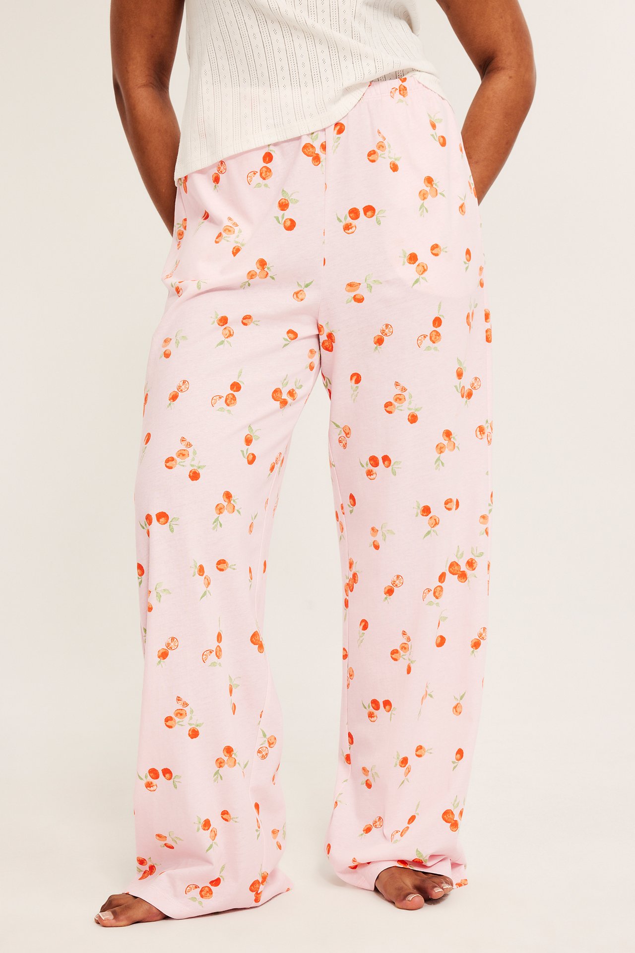 Edestä kuvattuna nainen yllään vaaleanpunaiset puuvillapyjamahousut, joissa oranssi hedelmäkuvio, ja valkoinen ribbitoppi.