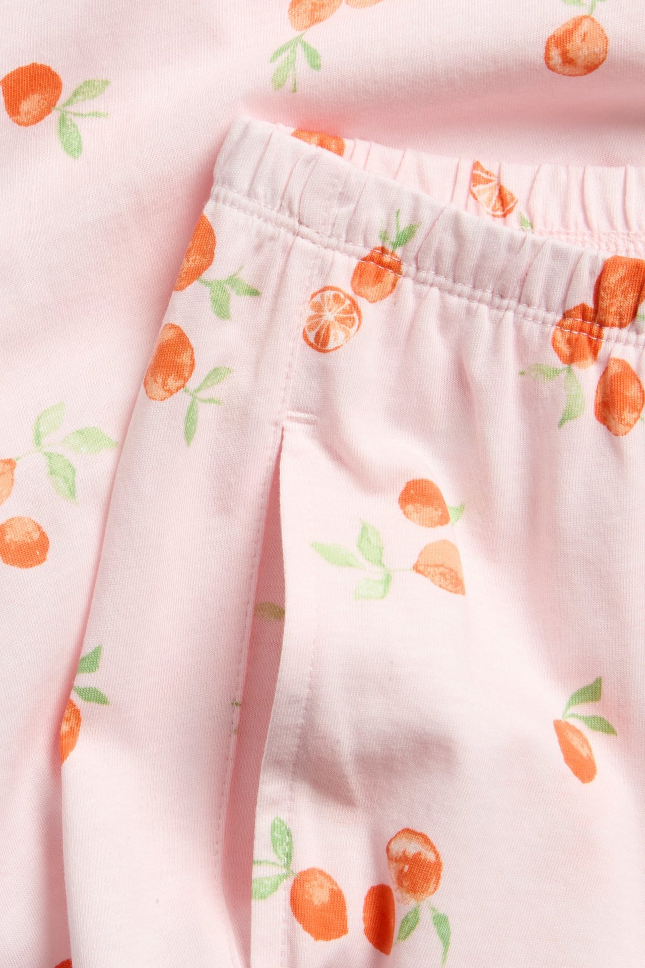 Lähikuva: vaaleanpunaiset trikoiset pyjamahousut oranssilla hedelmäkuviolla, joustava vyötärö ja sivutasku.