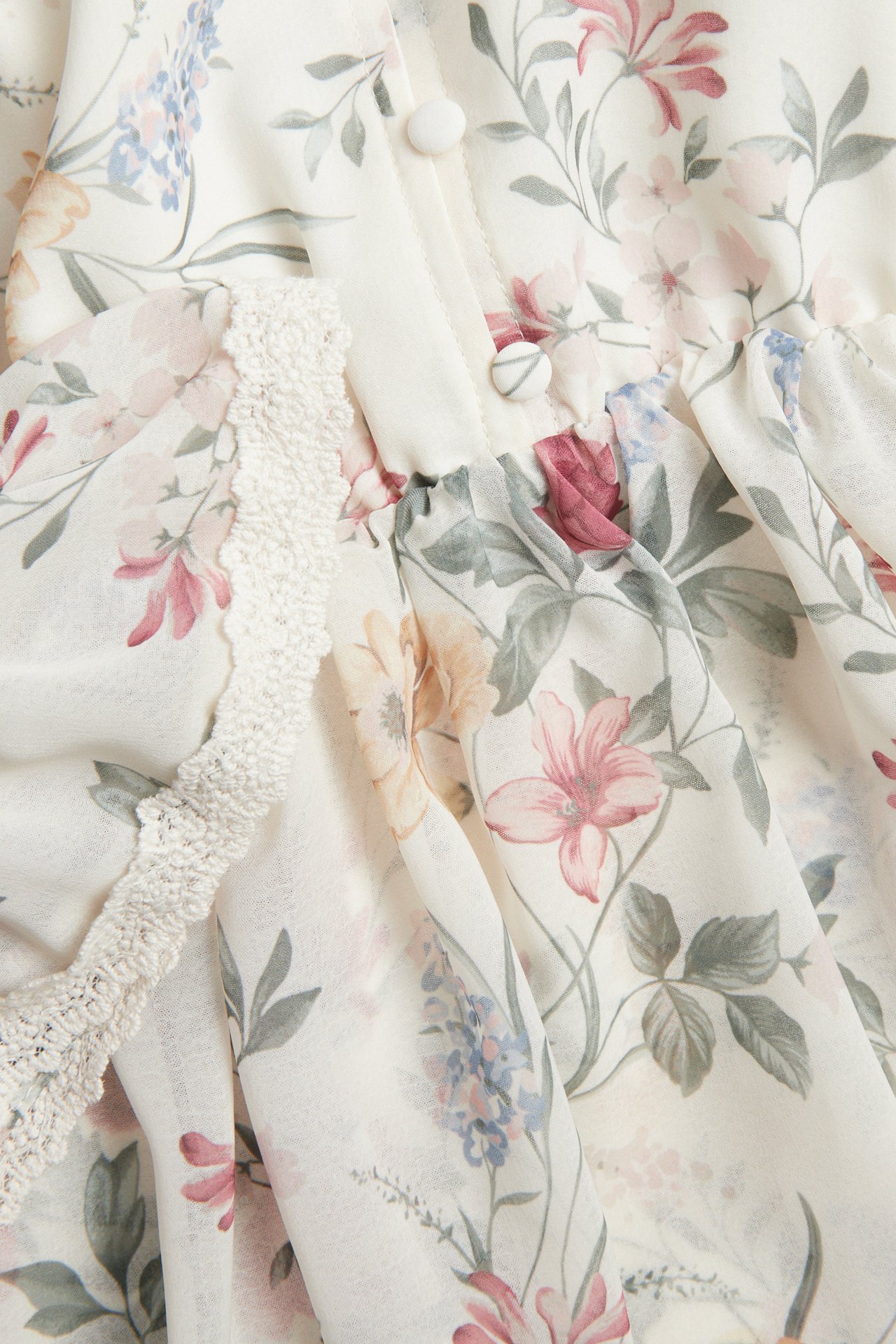 Nærbilde av en offwhite blomstret chiffonkjole med rosa, blå, grønne blomster og stofftrukne knapper.