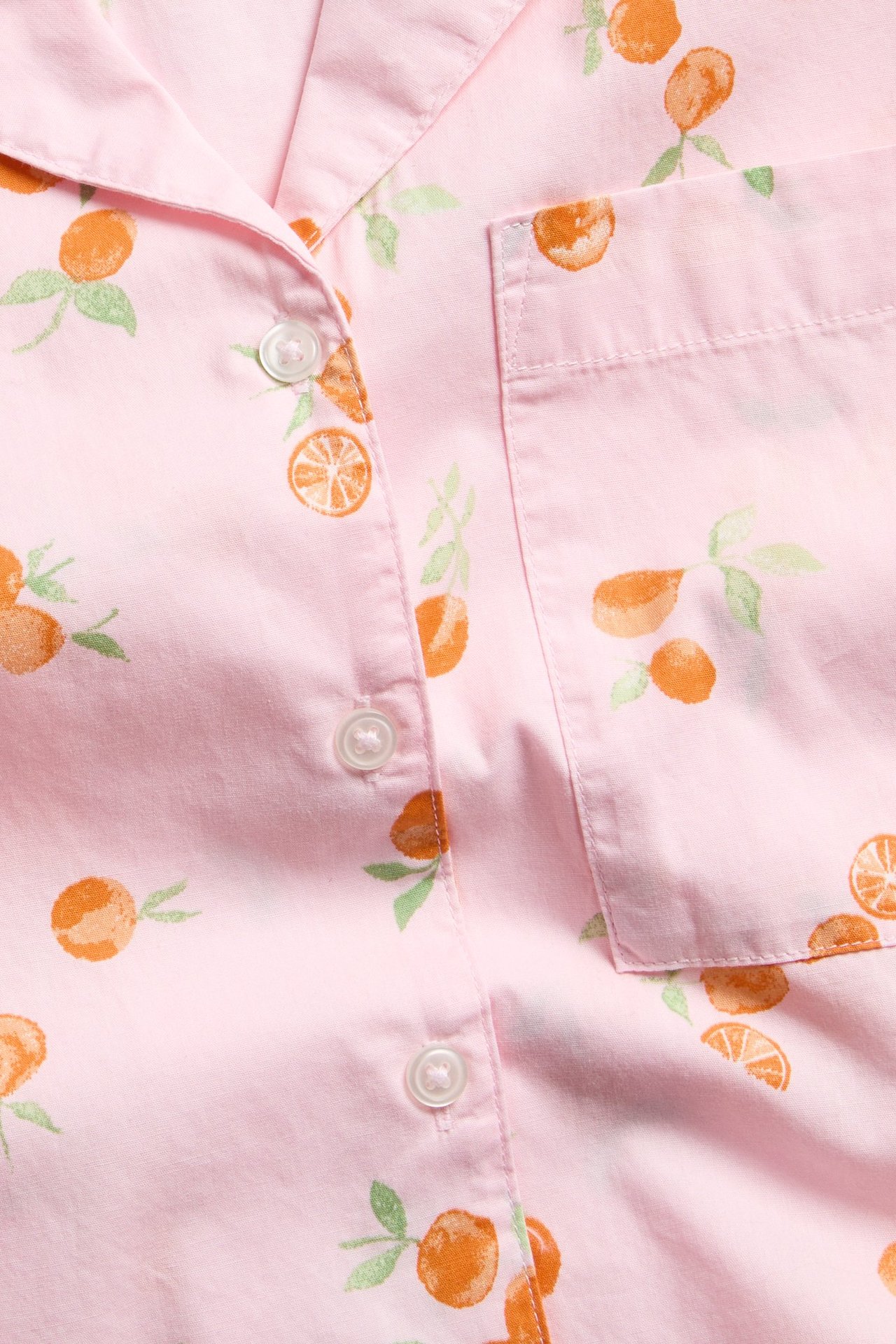 Zbliżenie jasnoróżowej koszuli piżamowej z tkanej bawełny z pomarańczowym wzorem, białymi guzikami i kieszenią.