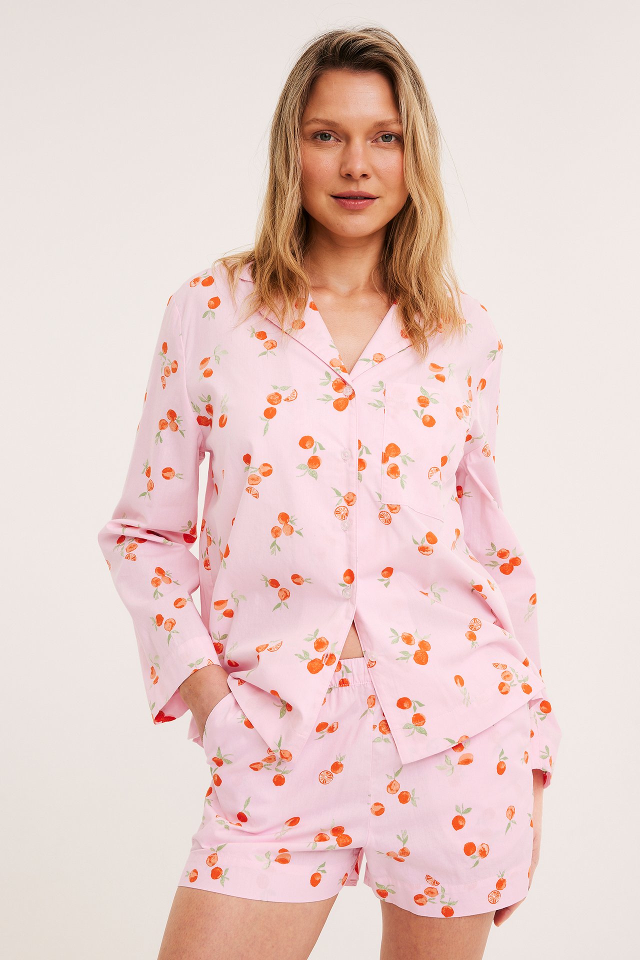 Etunäkymä: Nainen vaaleanpunaisissa puuvillapyjamashortseissa, joissa appelsiinikuvio, ja niihin sopiva nappipaita.