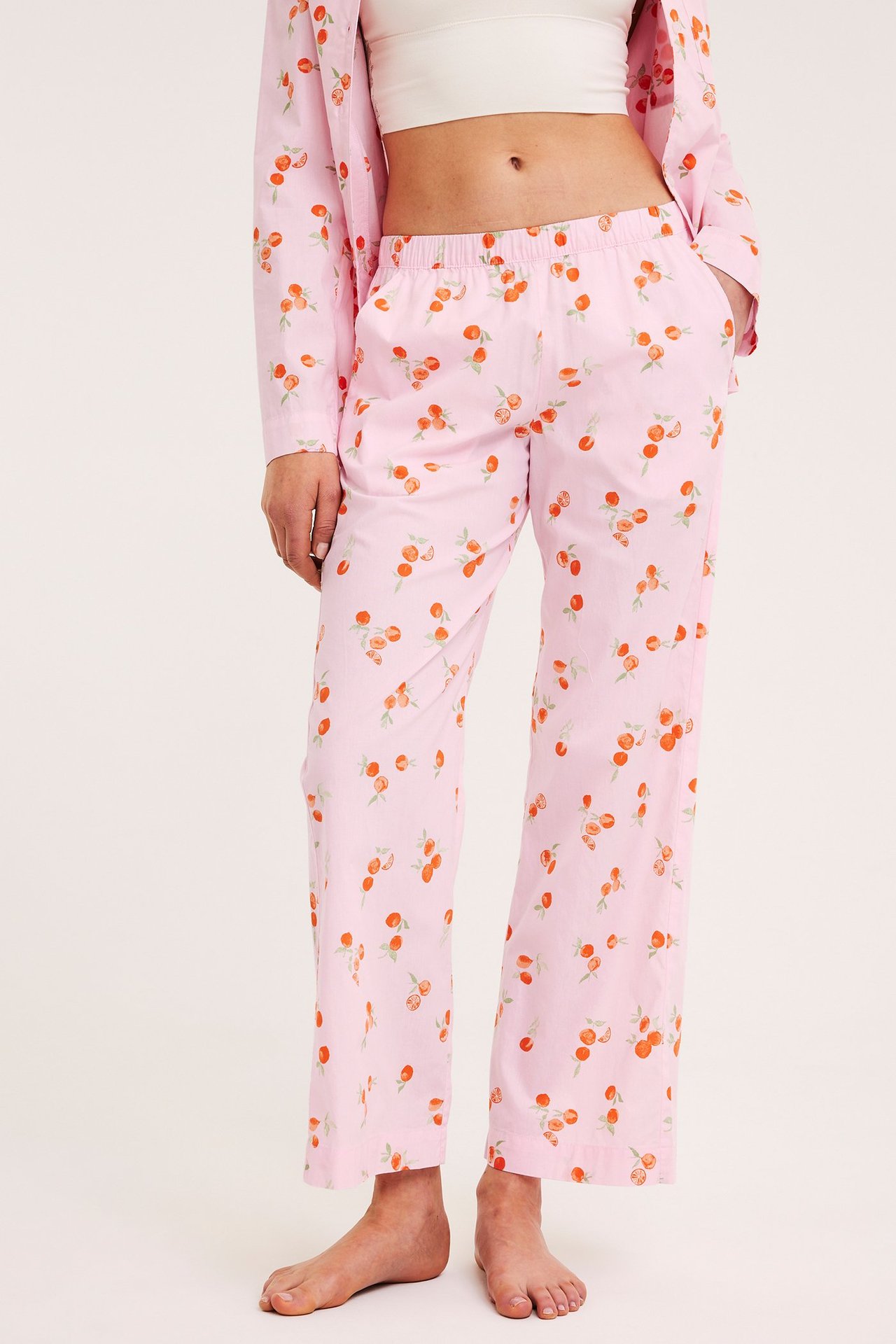 Nainen yllään vaaleanpunaiset hedelmäkuvioiset puuvillapyjamahousut, joustovyötärö, valkoinen toppi ja yhteensopiva paita.